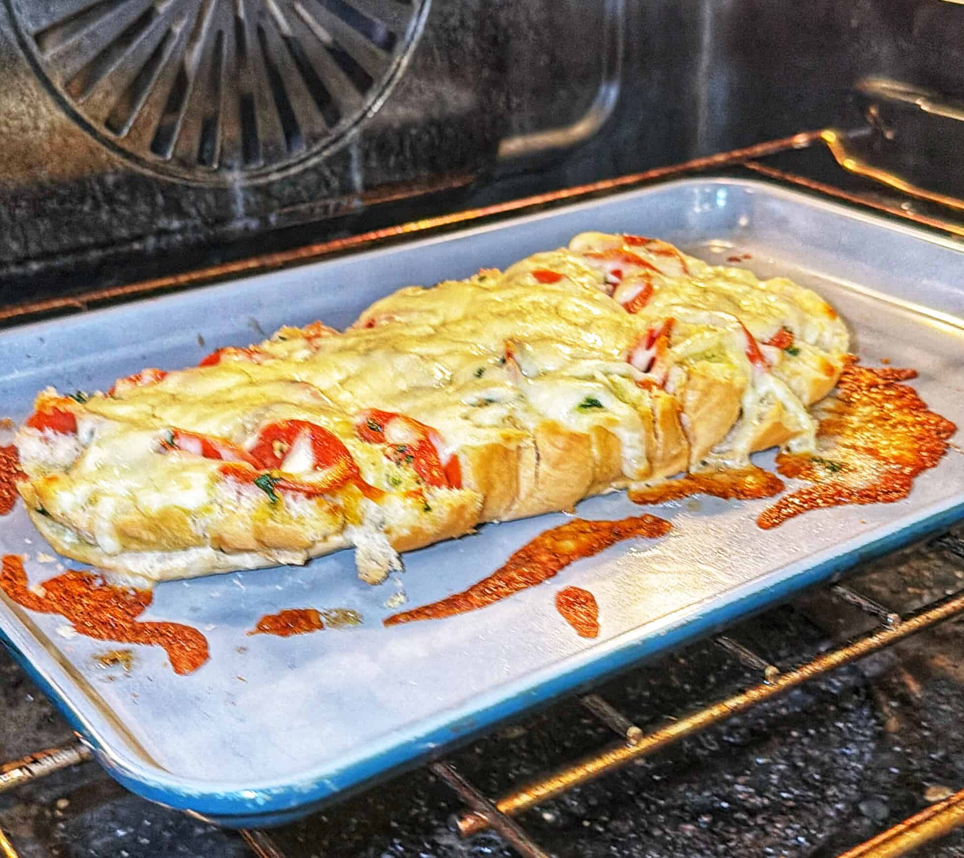 A loaf of bread topped with melted cheese, sliced tomatoes, and herbs is baking on a baking sheet in an oven. Some cheese has melted onto the pan.