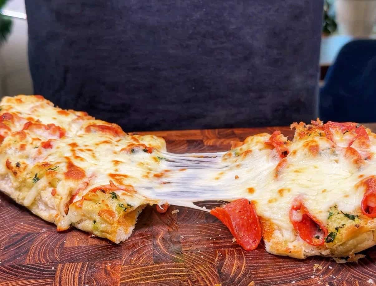 A close-up of cheesy pepperoni bread being pulled apart, showing melted cheese stretching between the pieces on a wooden board.
