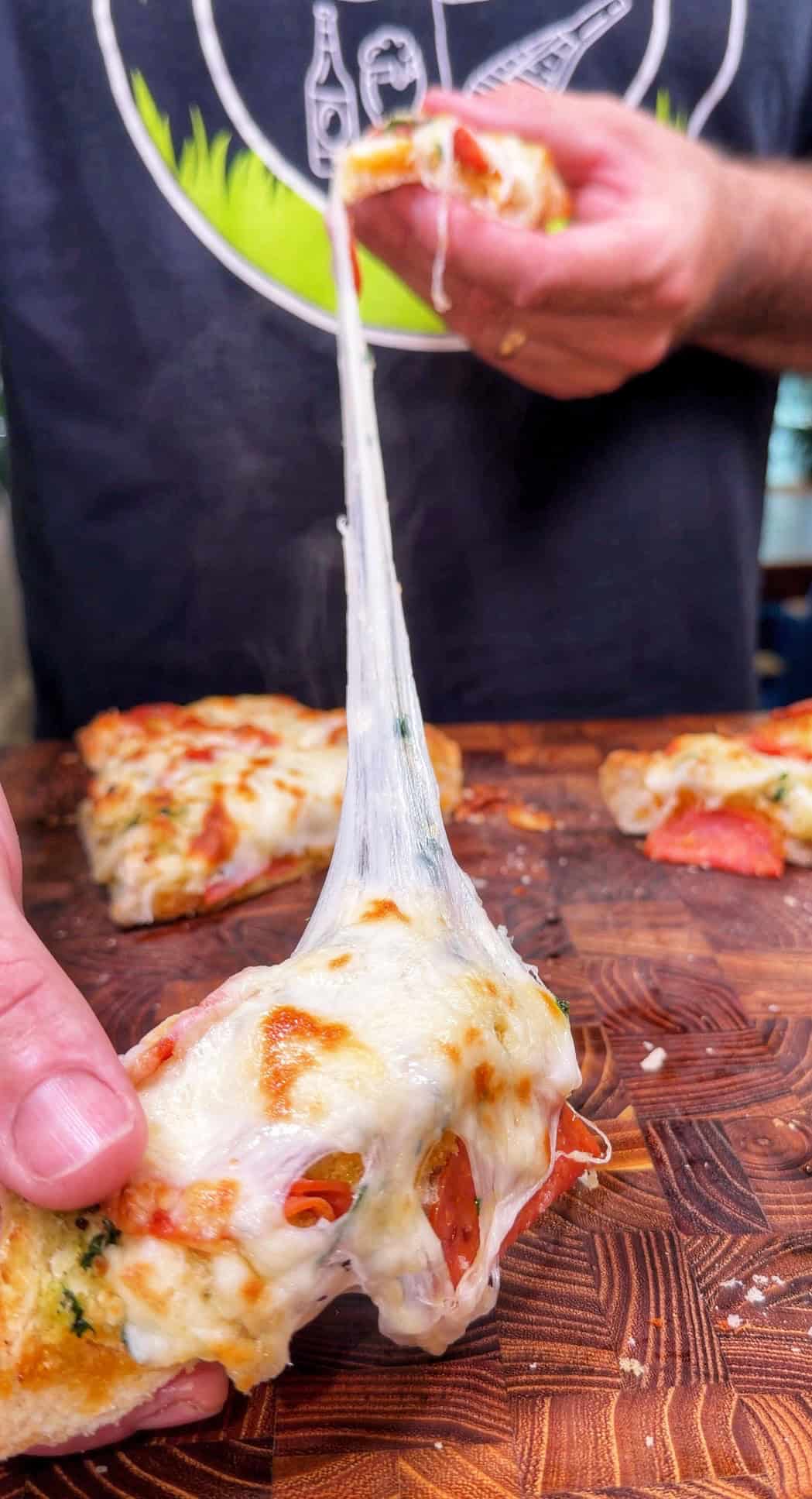 A hand lifts a cheesy slice of pizza from a wooden board, with melted cheese stretching between the slice and the remaining pizza.