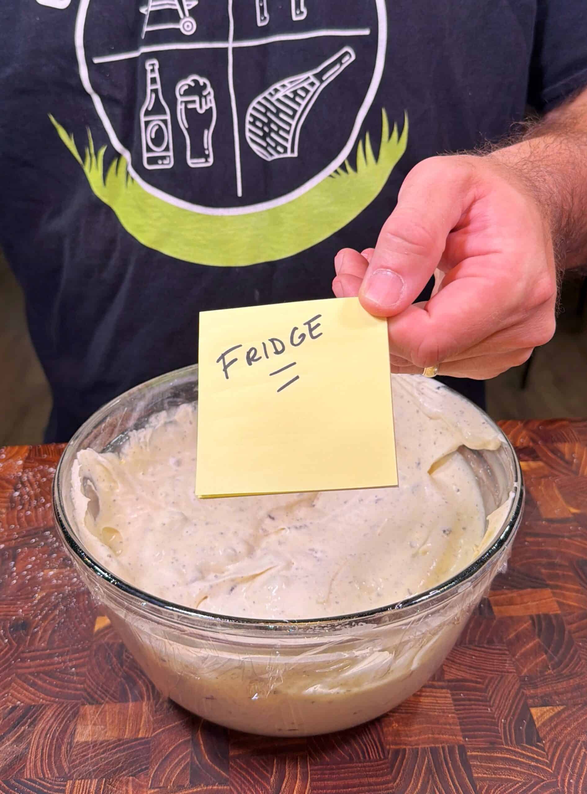 A person holds a yellow sticky note labeled FRIDGE over a glass bowl of batter on a wooden counter. The person is wearing a dark shirt with white cooking-themed graphics.