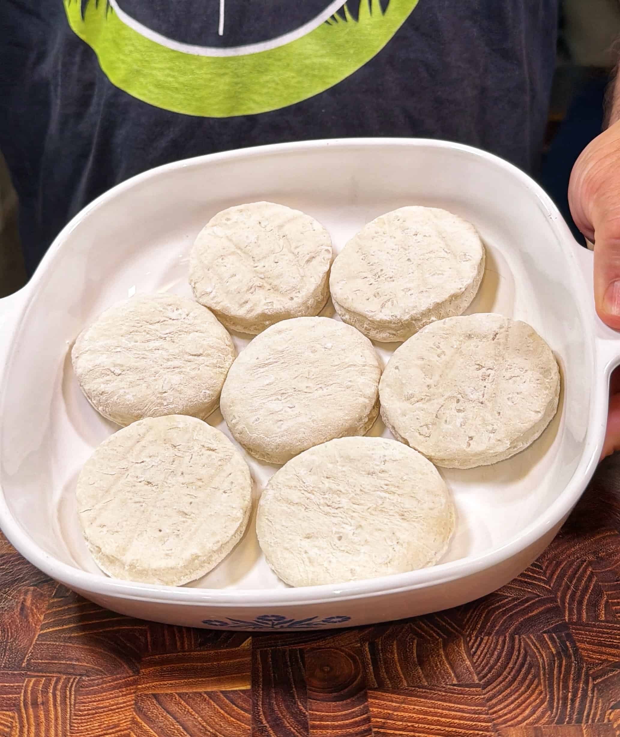 A person holds a white baking dish with seven uncooked, round biscuit dough pieces arranged inside. The dish is on a wooden surface.