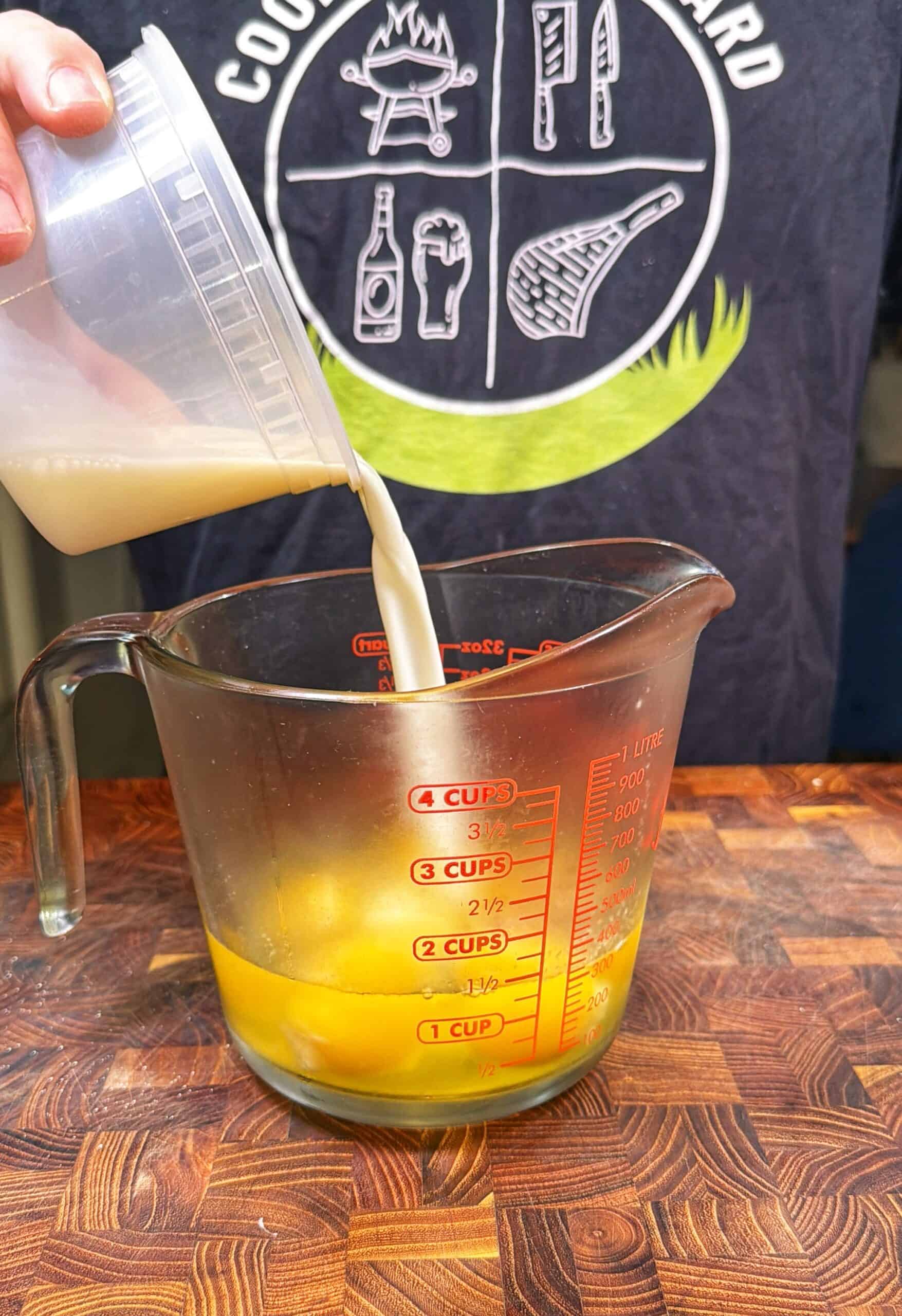 A person pours milk from a plastic container into a measuring cup containing cracked eggs on a wooden countertop. The person wears a dark shirt with white cooking-themed graphics.