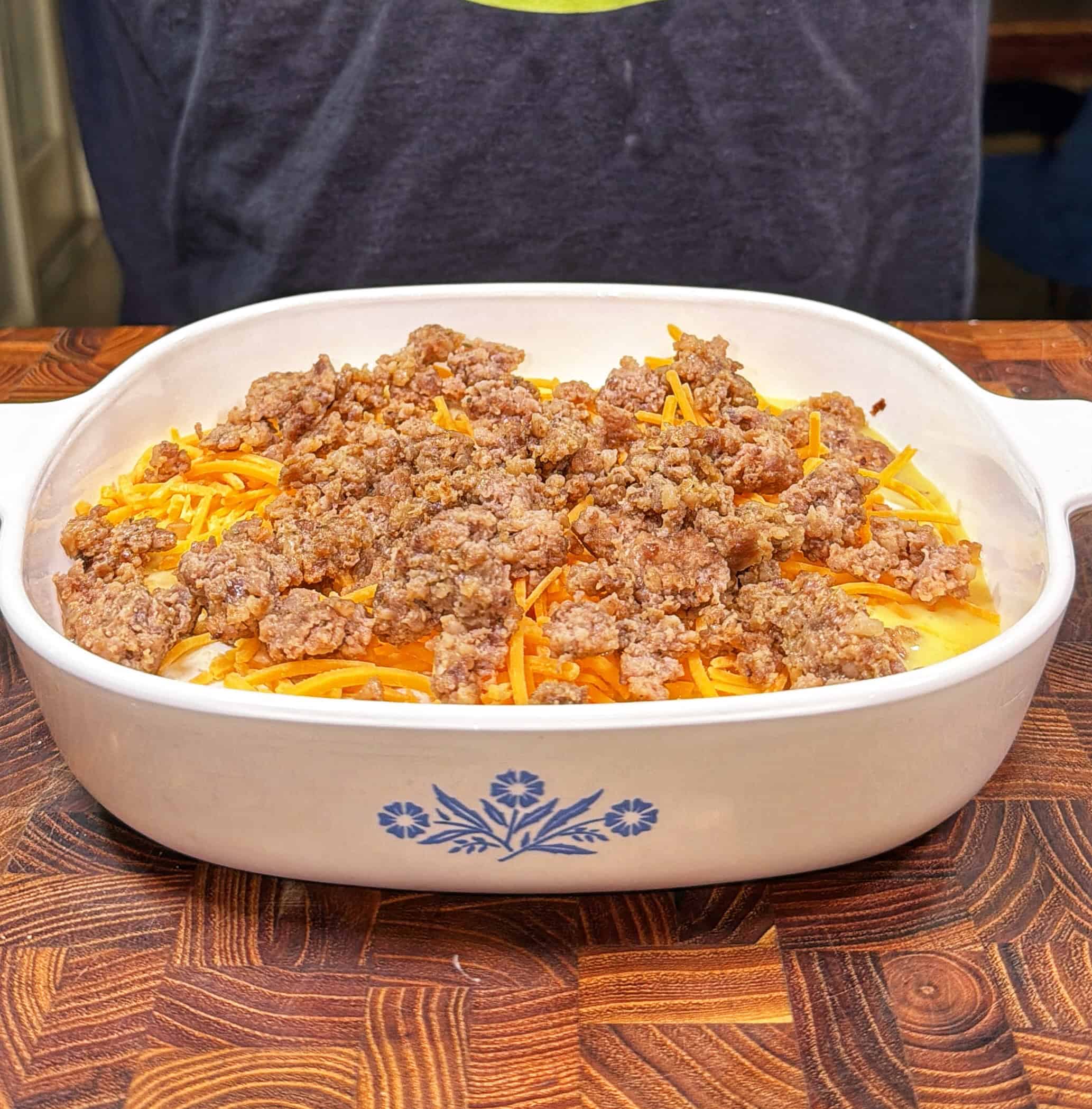 A white casserole dish filled with shredded cheddar cheese and cooked ground sausage sits on a wooden countertop, with a person in a dark shirt in the background.
