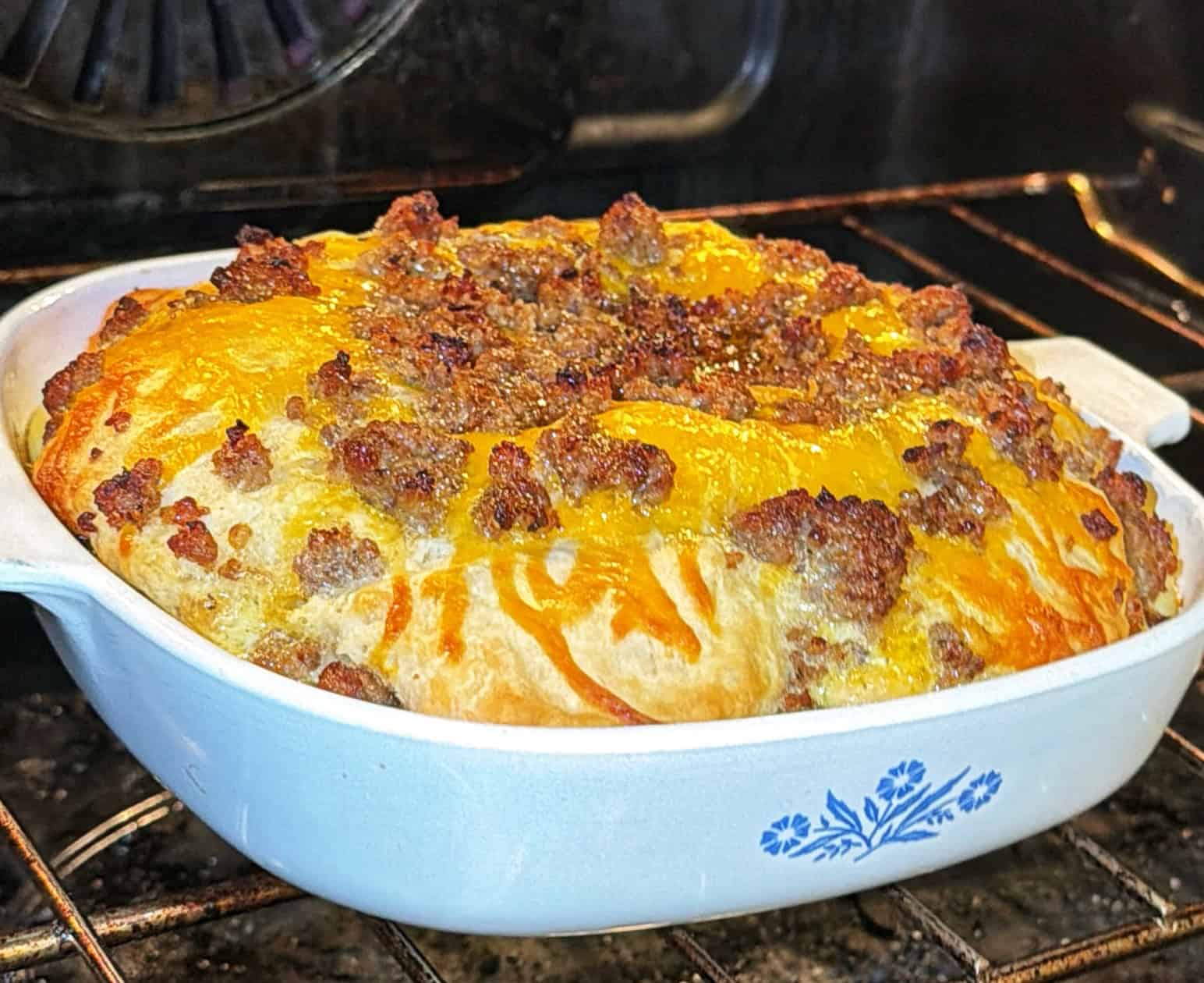 A breakfast casserole with melted cheese and browned sausage sits in a white baking dish with a blue floral design, fresh out of the oven.