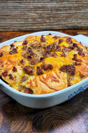 A baked casserole in a white dish, topped with melted cheese and crumbled sausage, sits on a wooden surface with a rustic wooden backdrop.