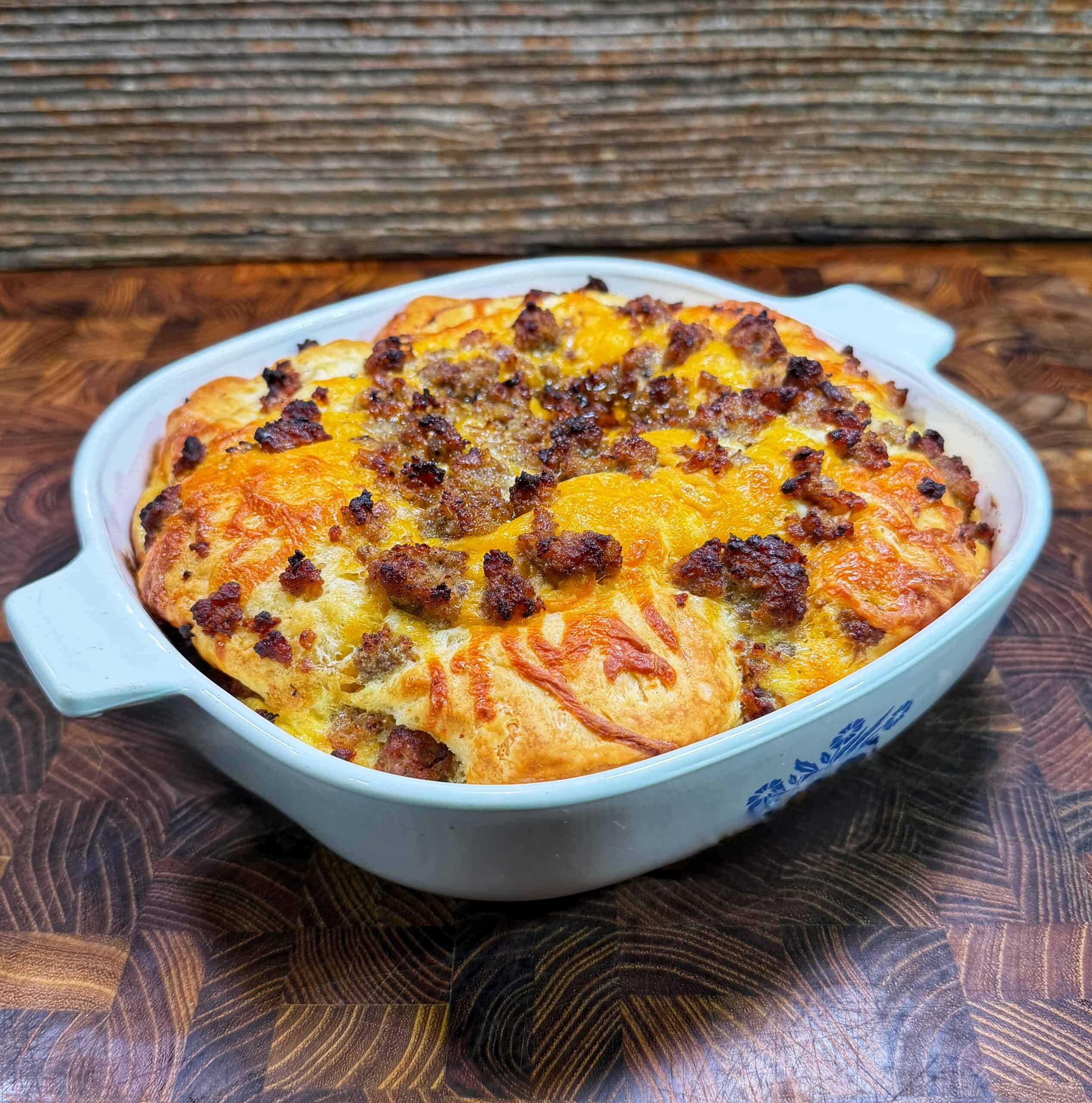 A baked casserole in a white dish, topped with melted cheese and crumbled sausage, sits on a wooden surface with a rustic wooden backdrop.