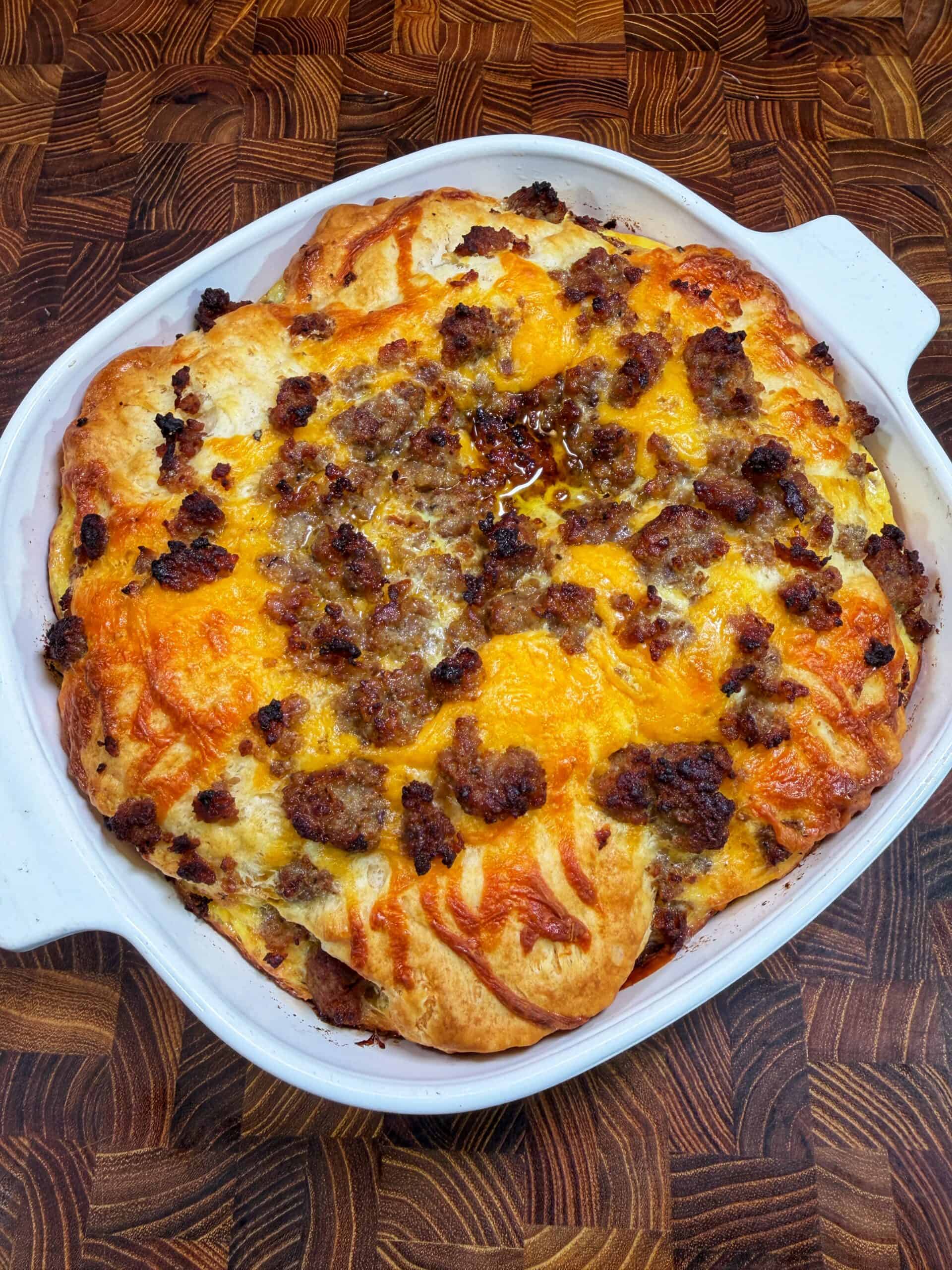 A baked casserole in a white square dish, topped with melted cheese and crumbled sausage, sits on a wooden countertop with a patterned surface.