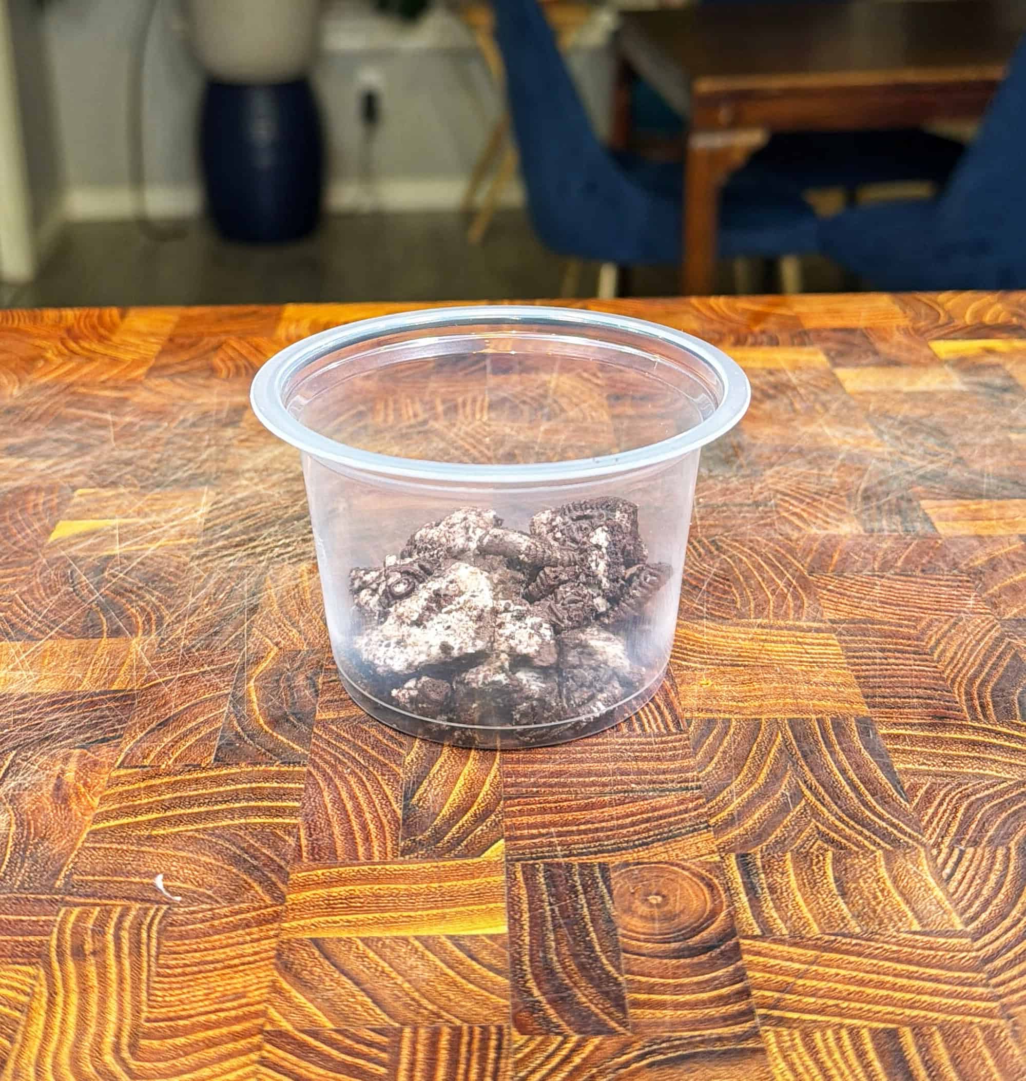 A small clear plastic container with chocolate cookie pieces sits on a wooden <a href=