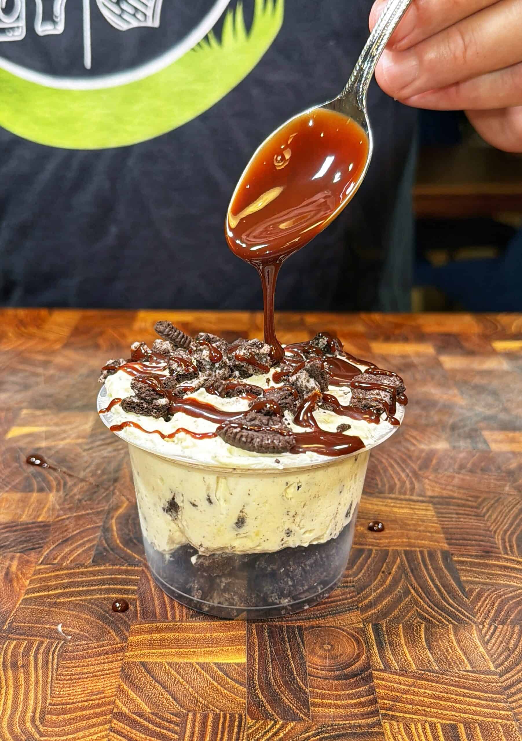 A hand drizzles chocolate sauce from a spoon onto a dessert cup filled with cookies and cream ice cream, topped with crumbled cookies, on a wooden surface.