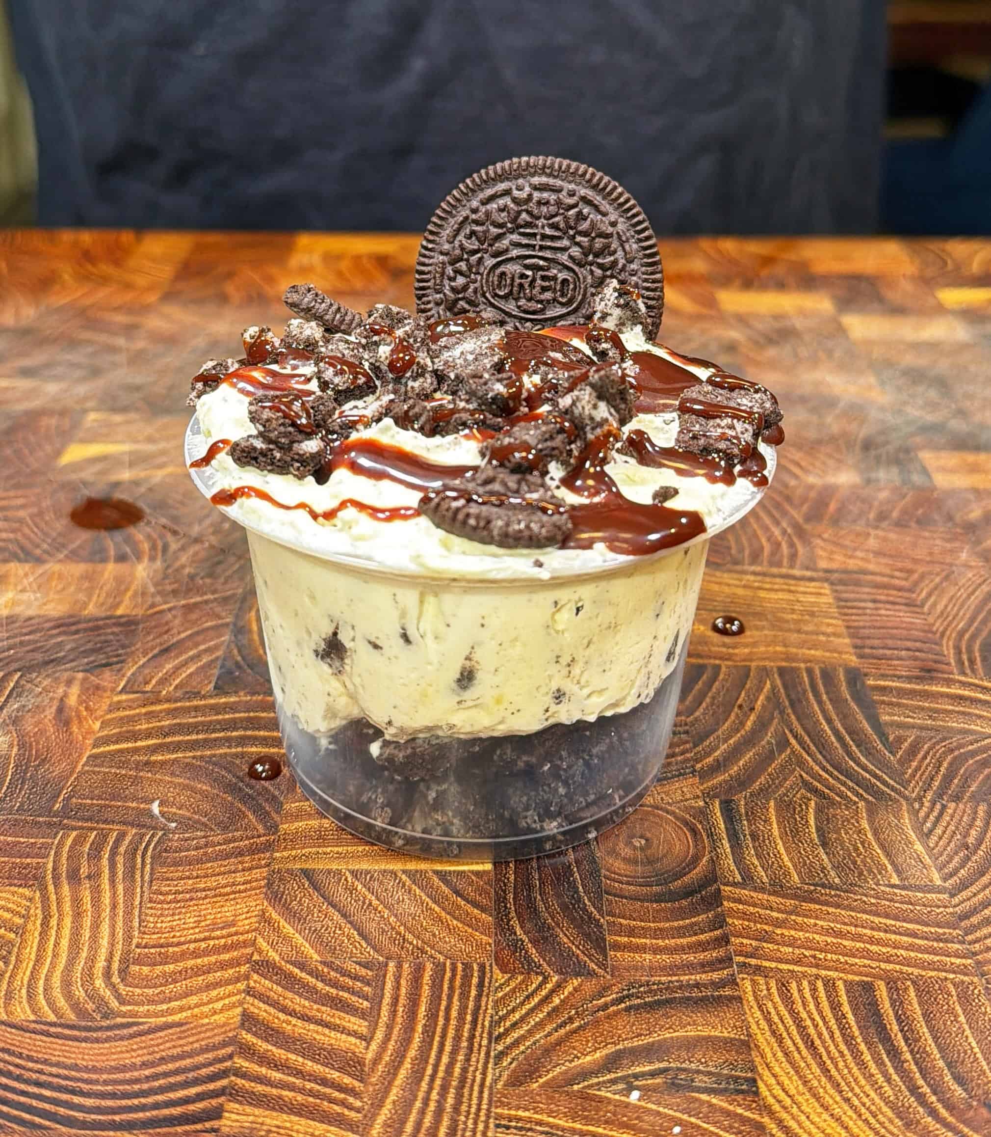A clear cup filled with cookies and cream dessert, topped with crushed cookies, chocolate drizzle, and a whole Oreo cookie, sits on a wooden surface.