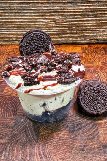 A cup of cookies and cream ice cream topped with crumbled Oreos and chocolate syrup, with a whole Oreo cookie on top and another Oreo resting on the wooden surface nearby.