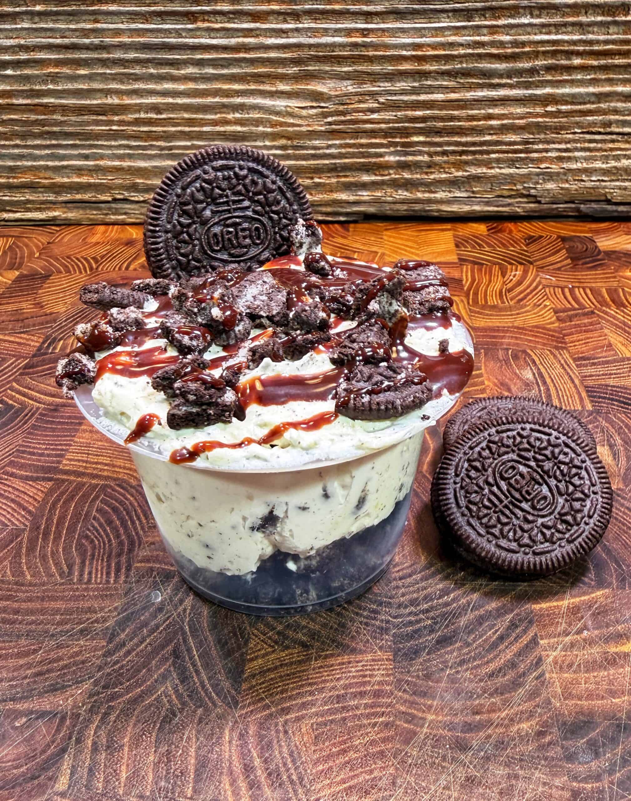 A cup of cookies and cream ice cream topped with crumbled Oreos and chocolate syrup, with a whole Oreo cookie on top and another Oreo resting on the wooden surface nearby.