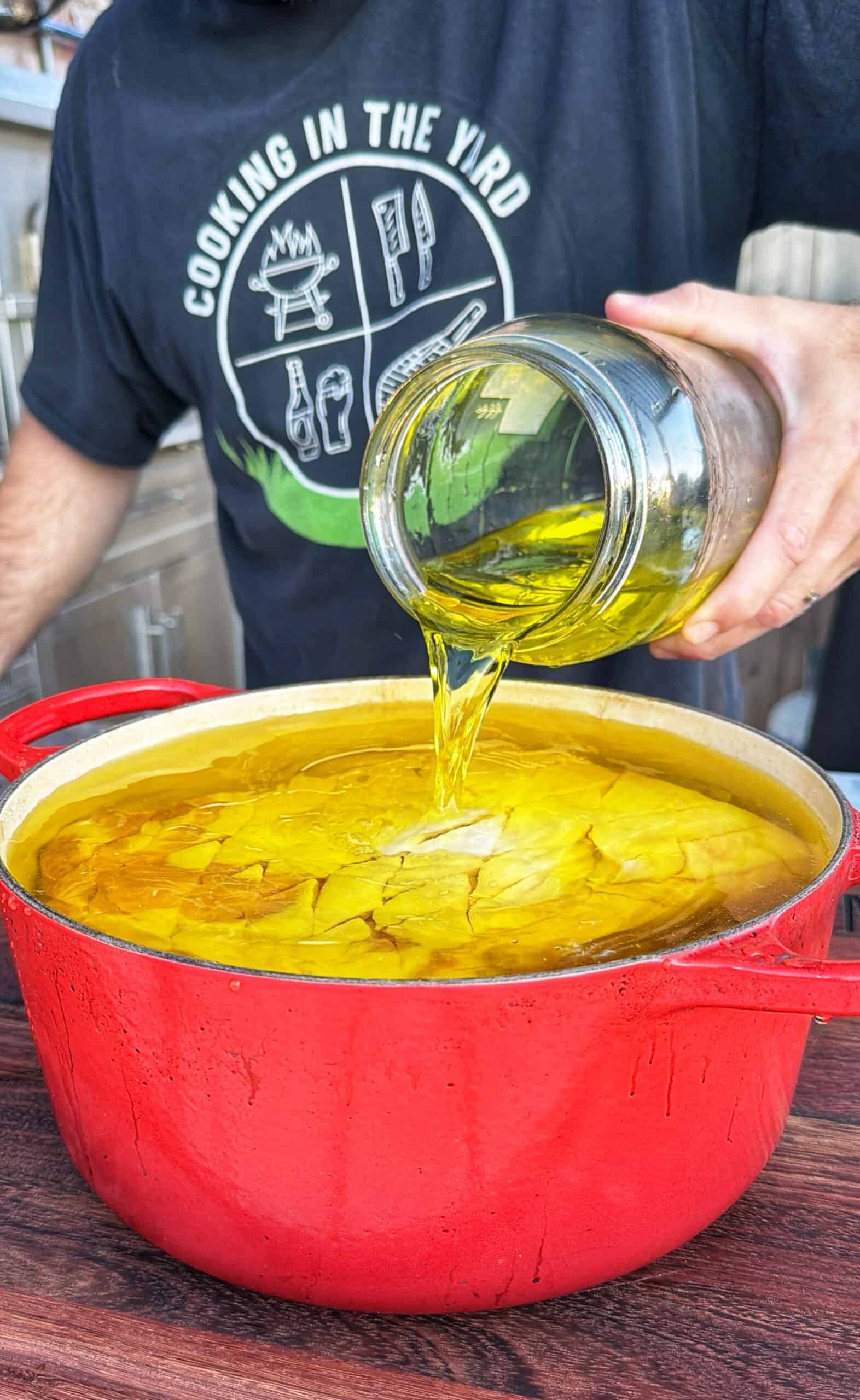 A person pours oil from a glass jar into a large red pot filled with oil. The person wears a black shirt that says COOKING IN THE YARD with cooking icons.