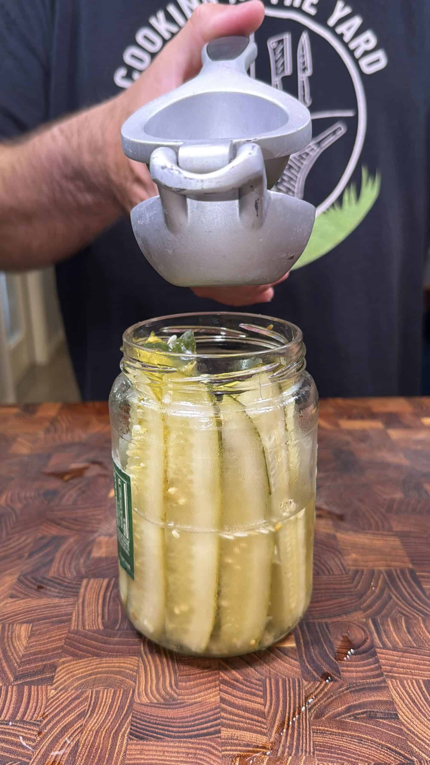 A hand squeezes a metal citrus press over a jar filled with pickle spears on a wooden surface.