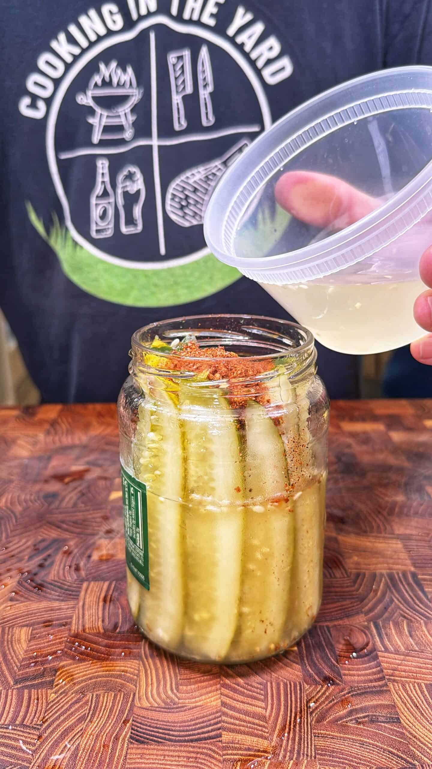 A hand pours liquid from a plastic lid into a jar of pickle spears with spices. A person wearing a Cooking in the Yard shirt stands behind the jar on a wooden surface.