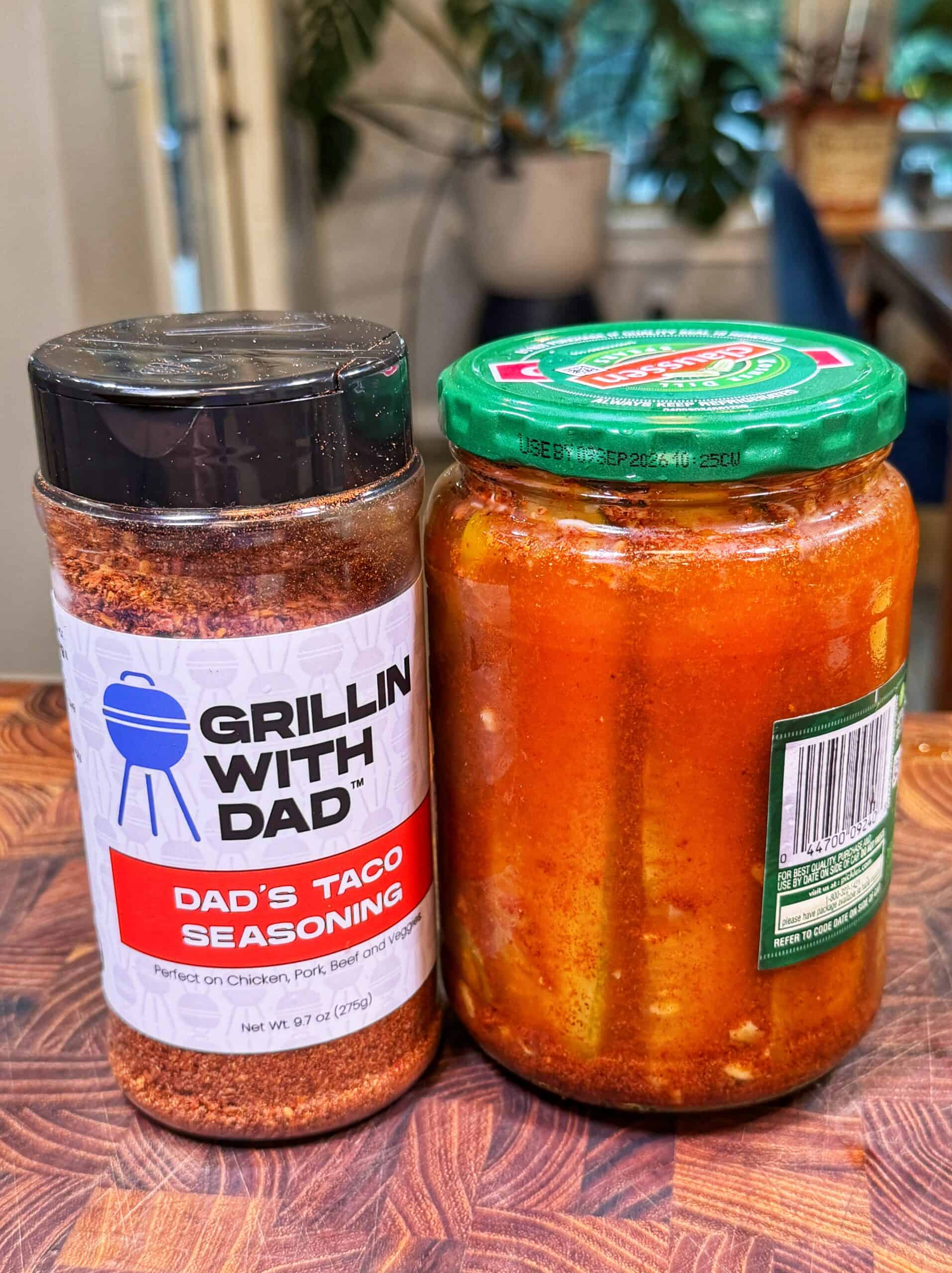 A jar of taco seasoning labeled Grillin With Dad next to a glass jar filled with red sauce on a wooden surface, with plants and blurred background indoors.