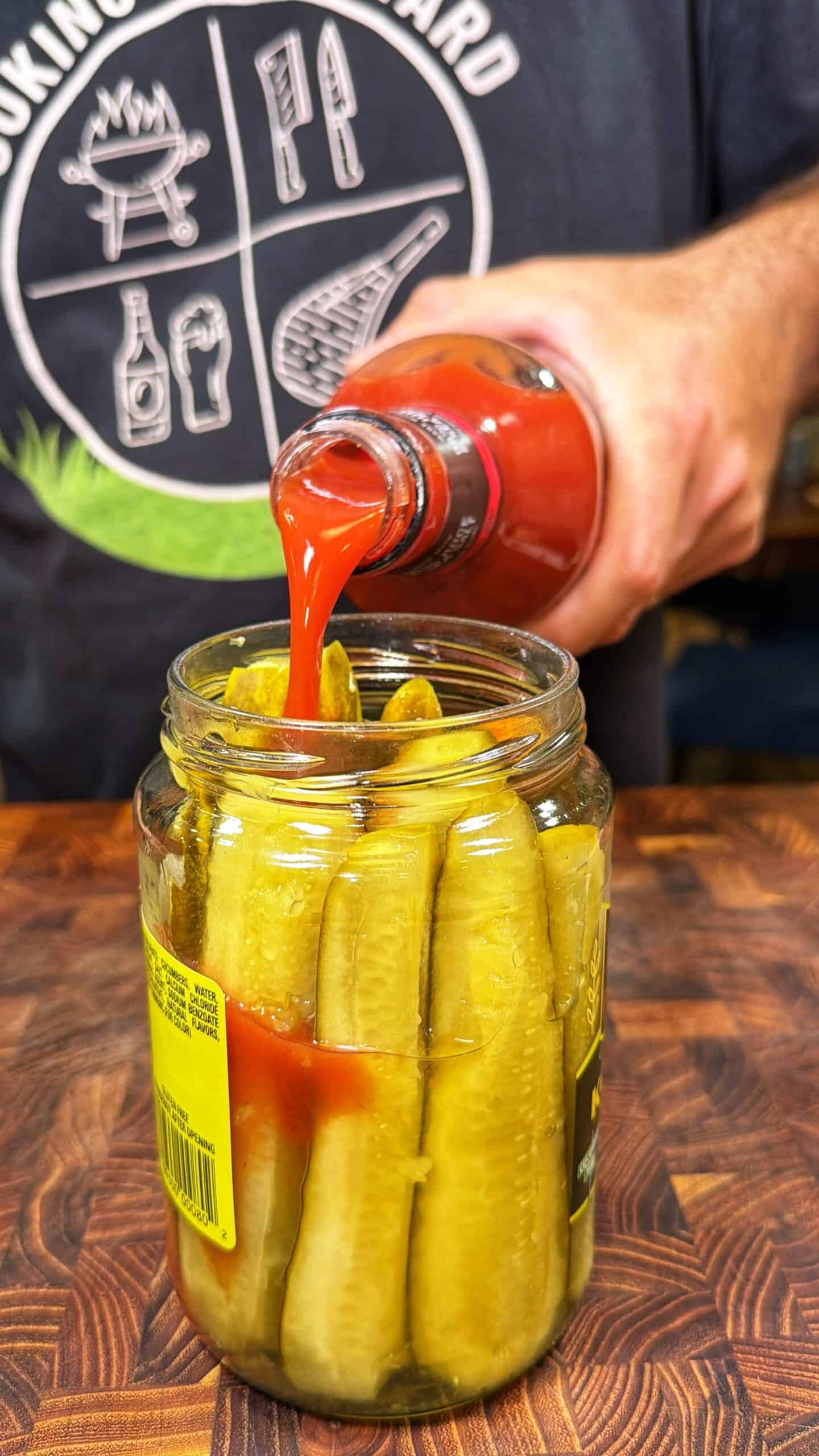 A person pours hot sauce from a bottle into a large jar of pickle spears on a wooden surface. The persons shirt has barbecue-themed graphics.