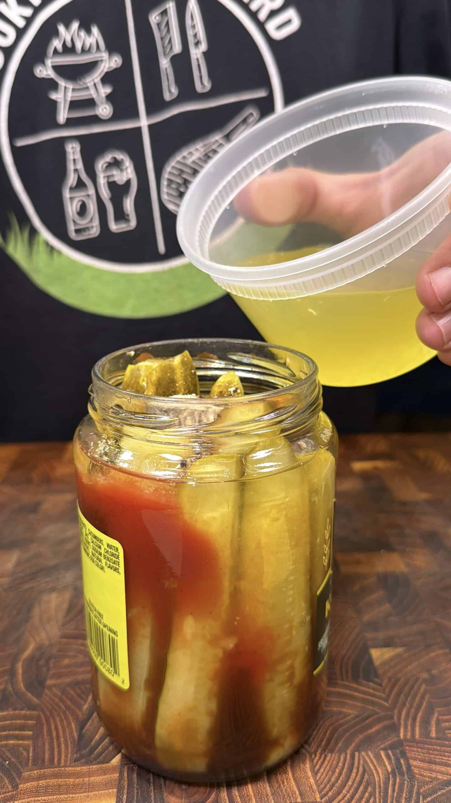 A hand pours yellow liquid from a plastic container into a jar of pickles with red sauce, on a wooden surface. A person wearing a black t-shirt with BBQ-themed graphics is in the background.