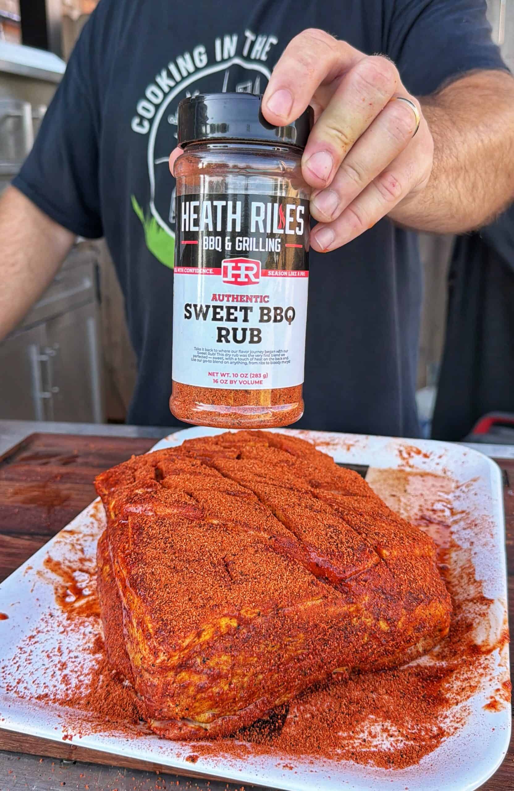 A hand holds a container of Heath Riles Authentic Sweet BBQ Rub above a plate with a large, seasoned piece of meat, ready for grilling. A person in a black shirt stands in the background.