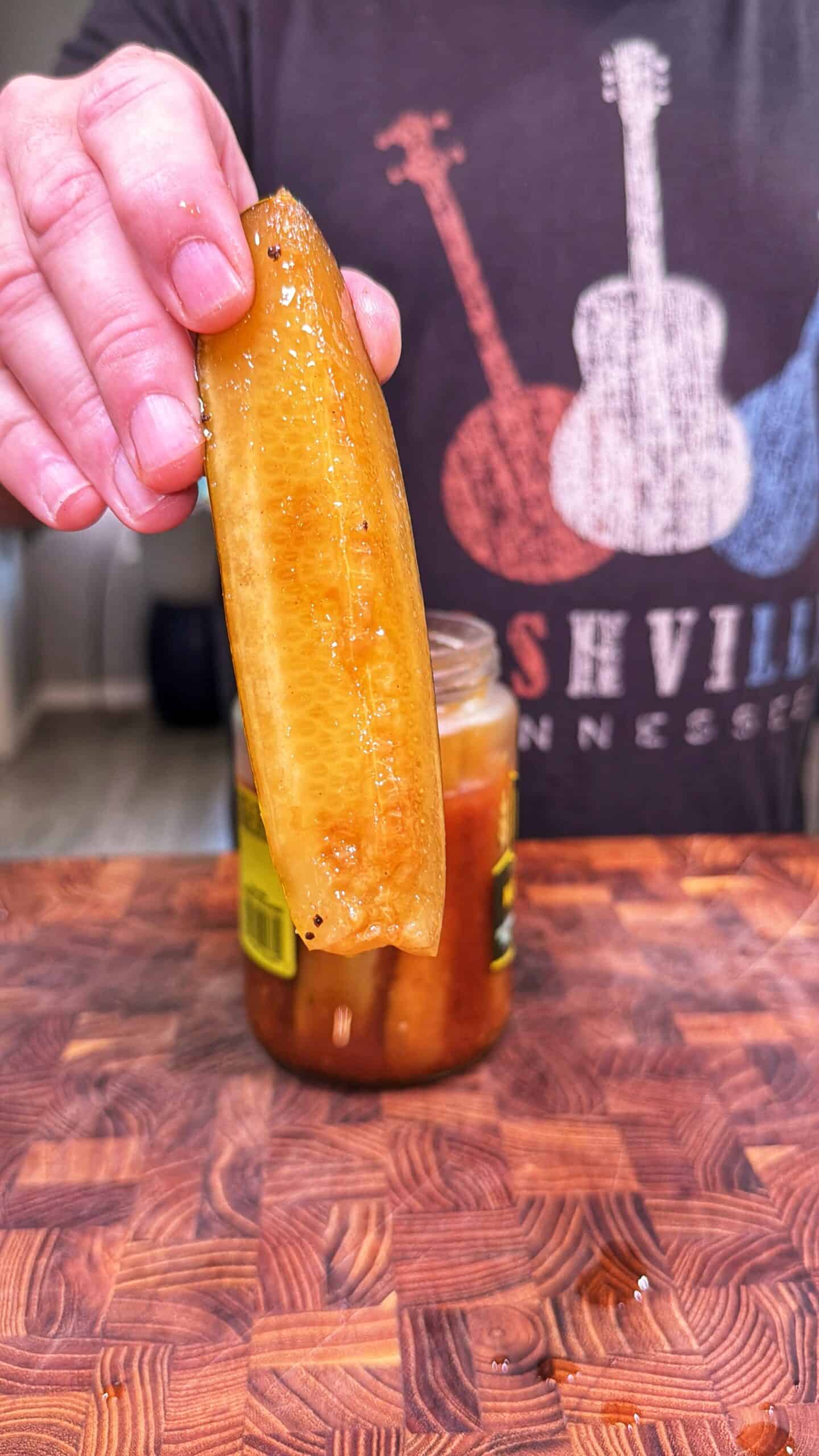 A person holding a pickle spear above a jar, with a wooden <a href=
