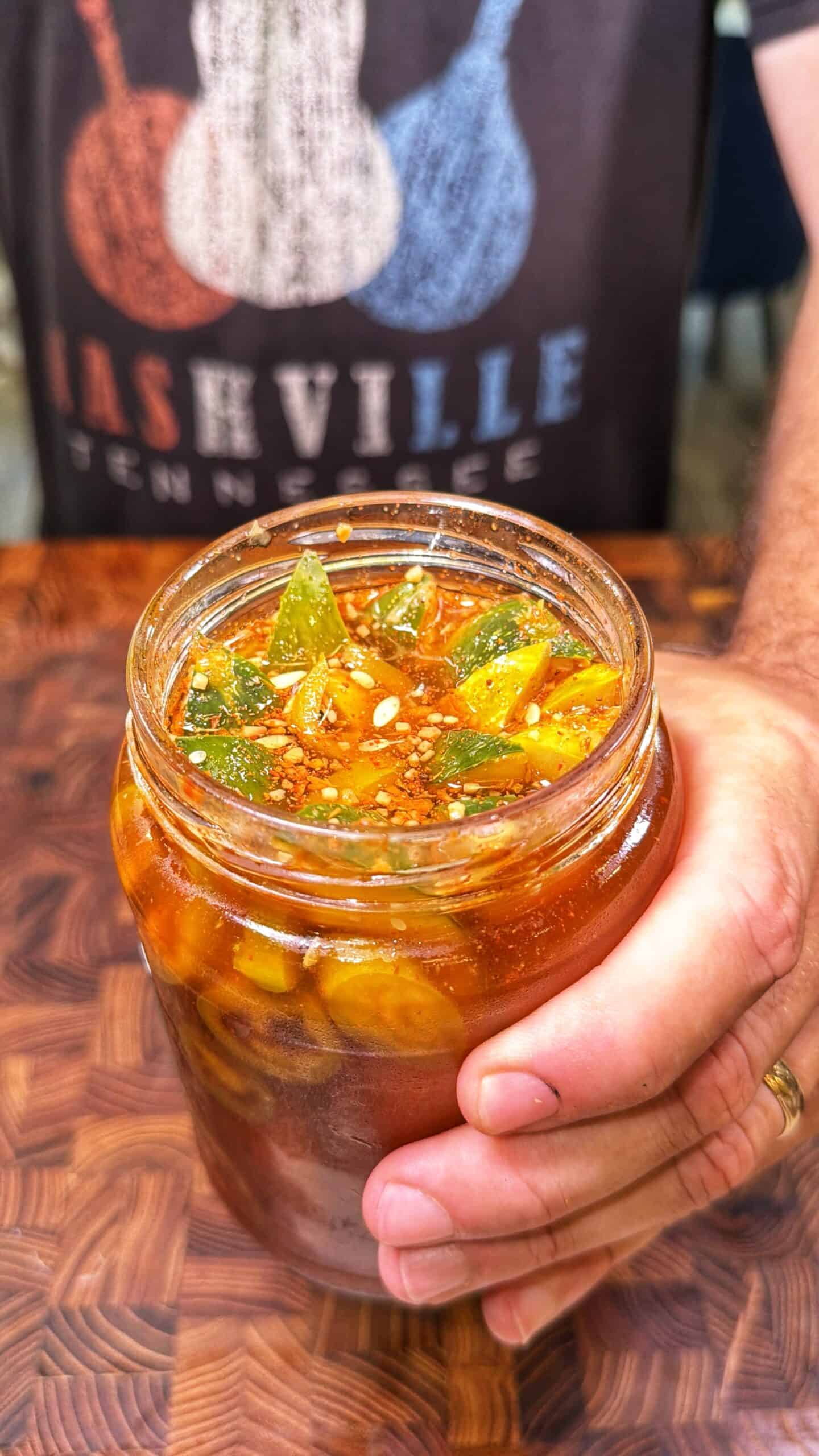A hand holds a glass jar filled with pickled vegetables in a spicy red brine. The background shows a person wearing a Nashville, Tennessee t-shirt.