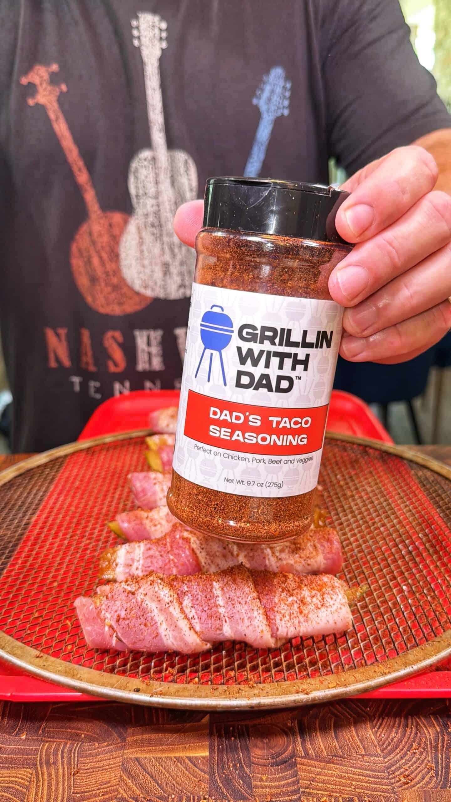 A person holds a bottle of Grillin With Dad Dads Taco Seasoning over raw bacon-wrapped food on a red wire rack. The person is wearing a Nashville shirt with guitar graphics.