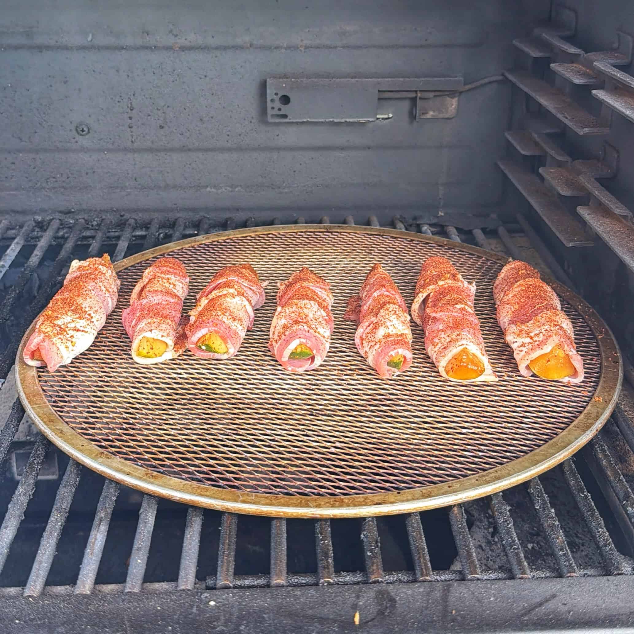 Six bacon-wrapped jalapeño poppers sit evenly spaced on a round mesh tray inside a grill, ready to be cooked.
