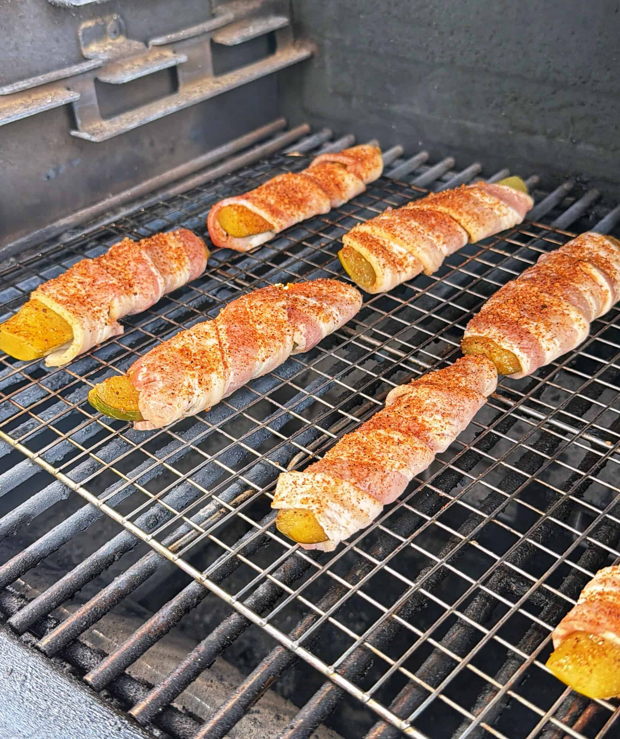 Pickles wrapped in bacon and seasoned with spices are arranged on a wire rack inside a barbecue grill, being cooked.