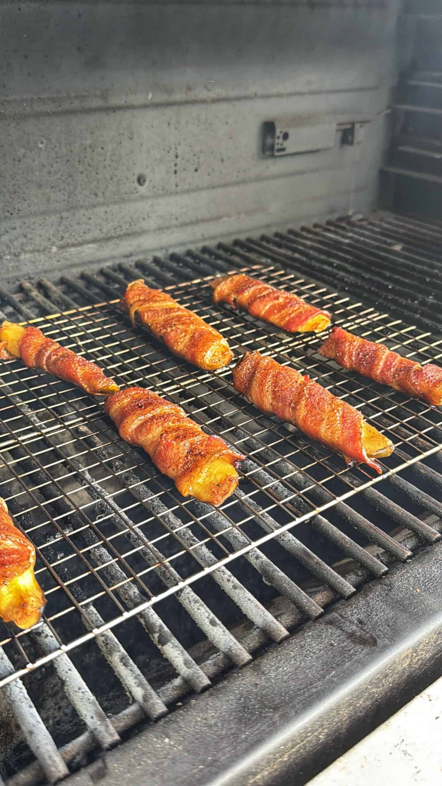 Six bacon-wrapped cheese sticks are arranged on a grill rack inside a barbecue grill, cooking with visible seasoning and grill marks.