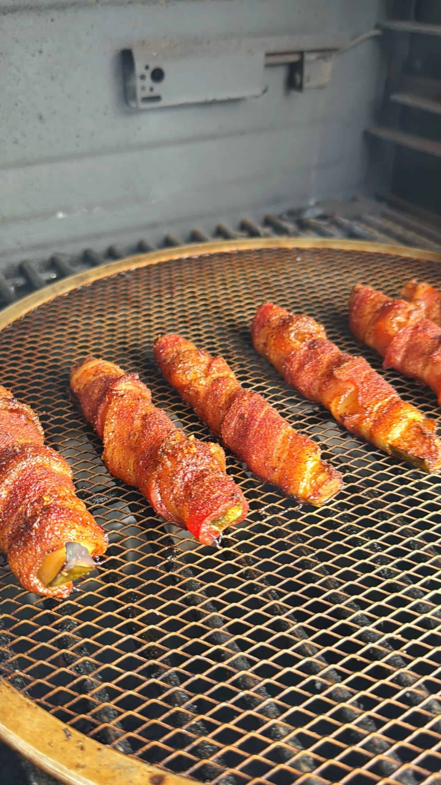 Several bacon-wrapped items are cooking on a metal grill inside a smoker, with a brown mesh grilling surface and visible seasoning on the bacon.
