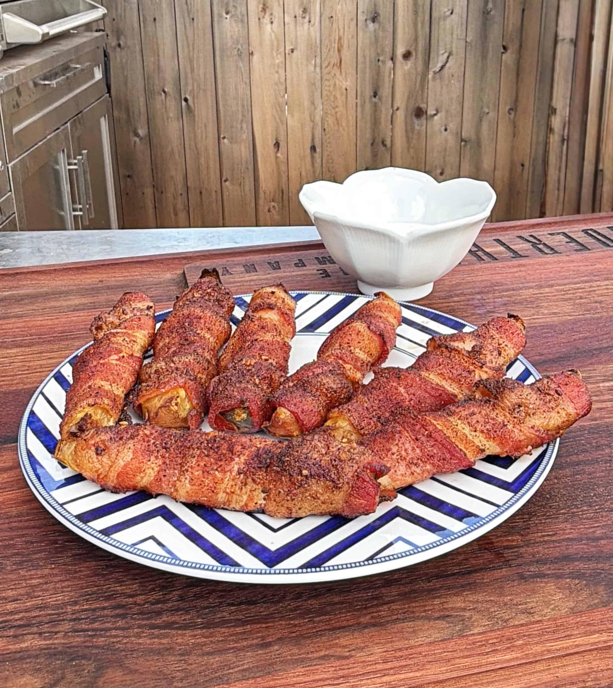 A plate of bacon-wrapped jalapeño poppers sits on a wooden table outdoors, with a white bowl in the background. The poppers are arranged neatly on a blue and white patterned plate.