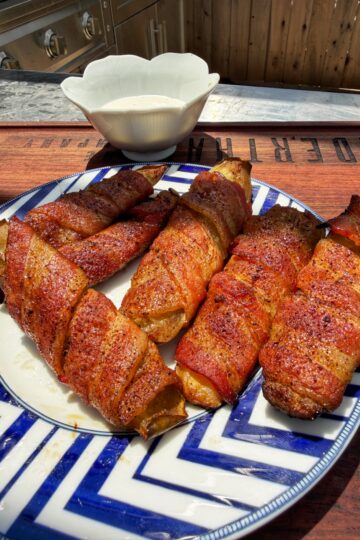 A blue and white plate holds several bacon-wrapped, grilled food items, seasoned and arranged outdoors on a wooden surface. A white dipping bowl sits in the background.