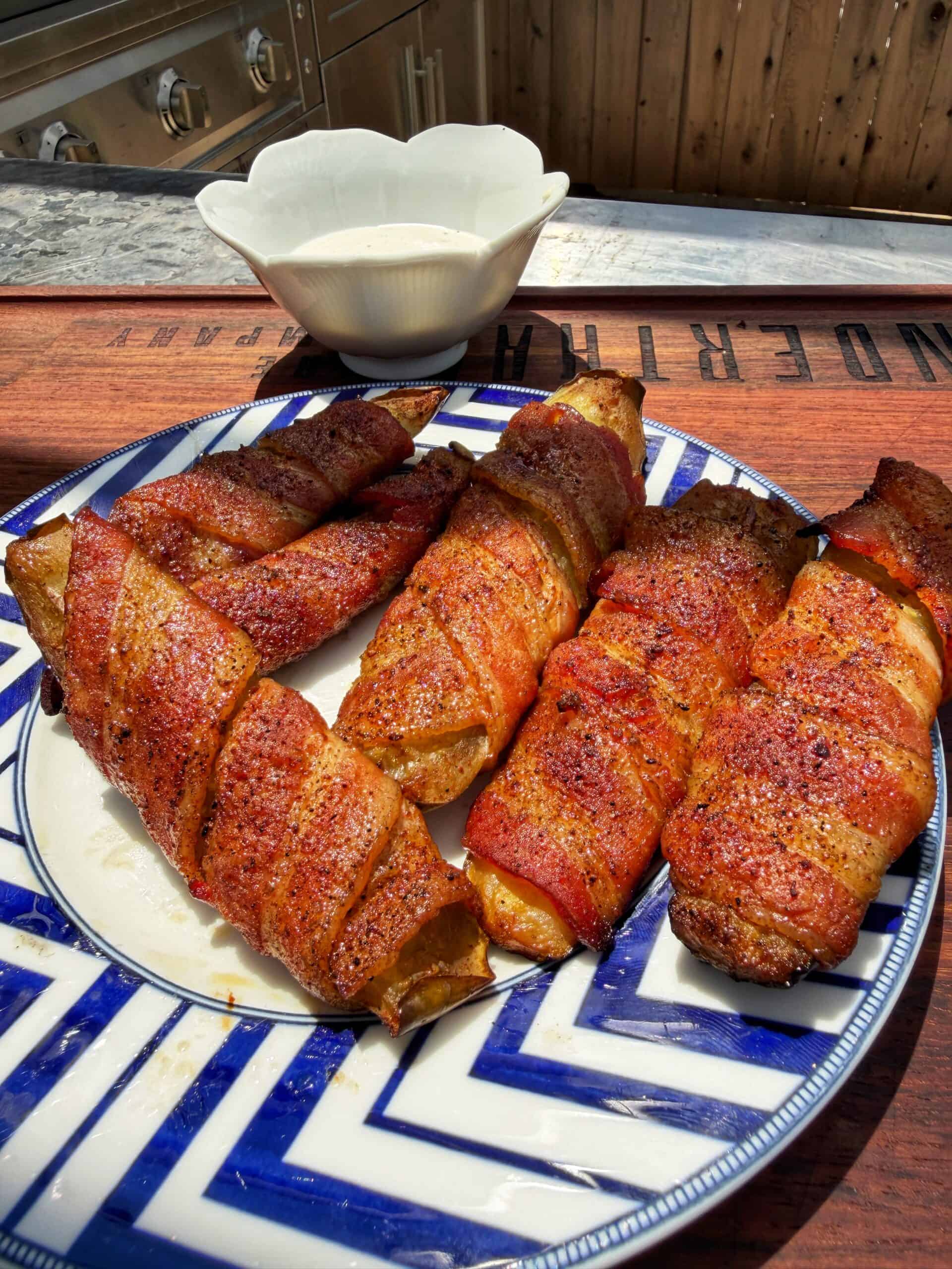 A blue and white plate holds several bacon-wrapped, grilled food items, seasoned and arranged outdoors on a wooden surface. A white dipping bowl sits in the background.