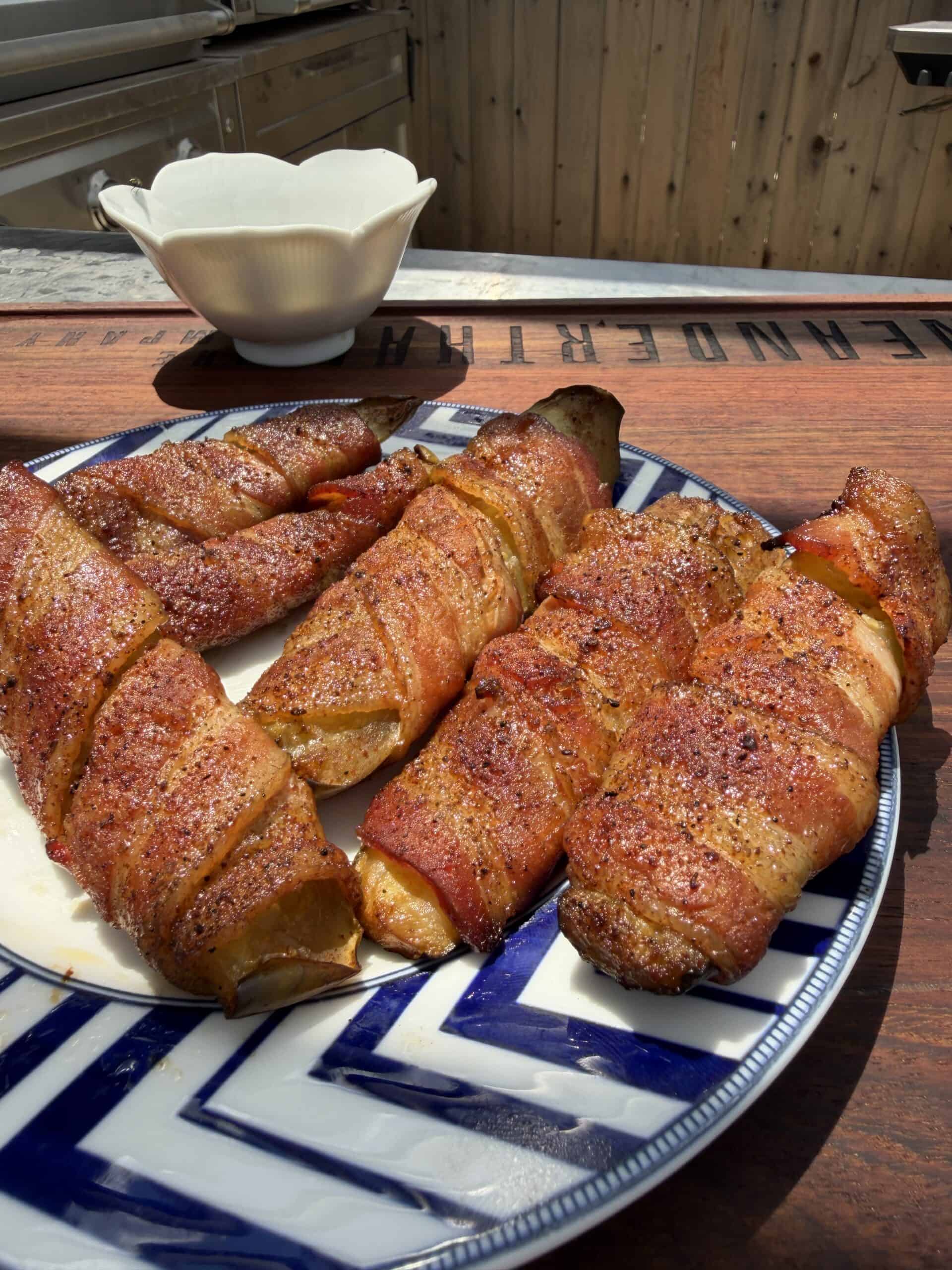 Four bacon-wrapped grilled items are arranged on a blue and white plate, sitting on a wooden surface outdoors, with a white bowl and wooden fence in the background.