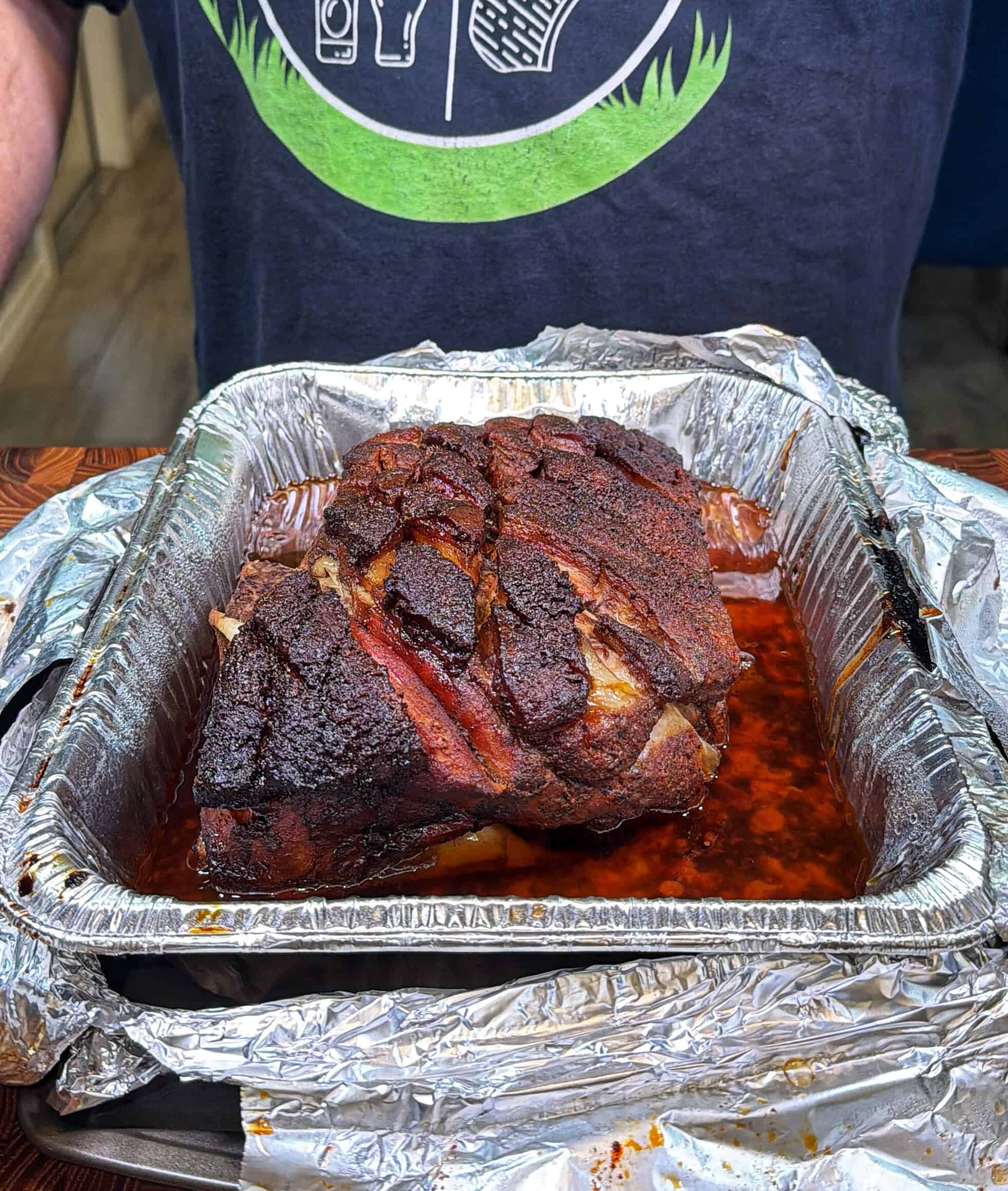 A large piece of smoked, seasoned meat sits in a foil-lined pan with juices, set on a table. A person wearing a dark shirt with a grill graphic is visible behind the pan.