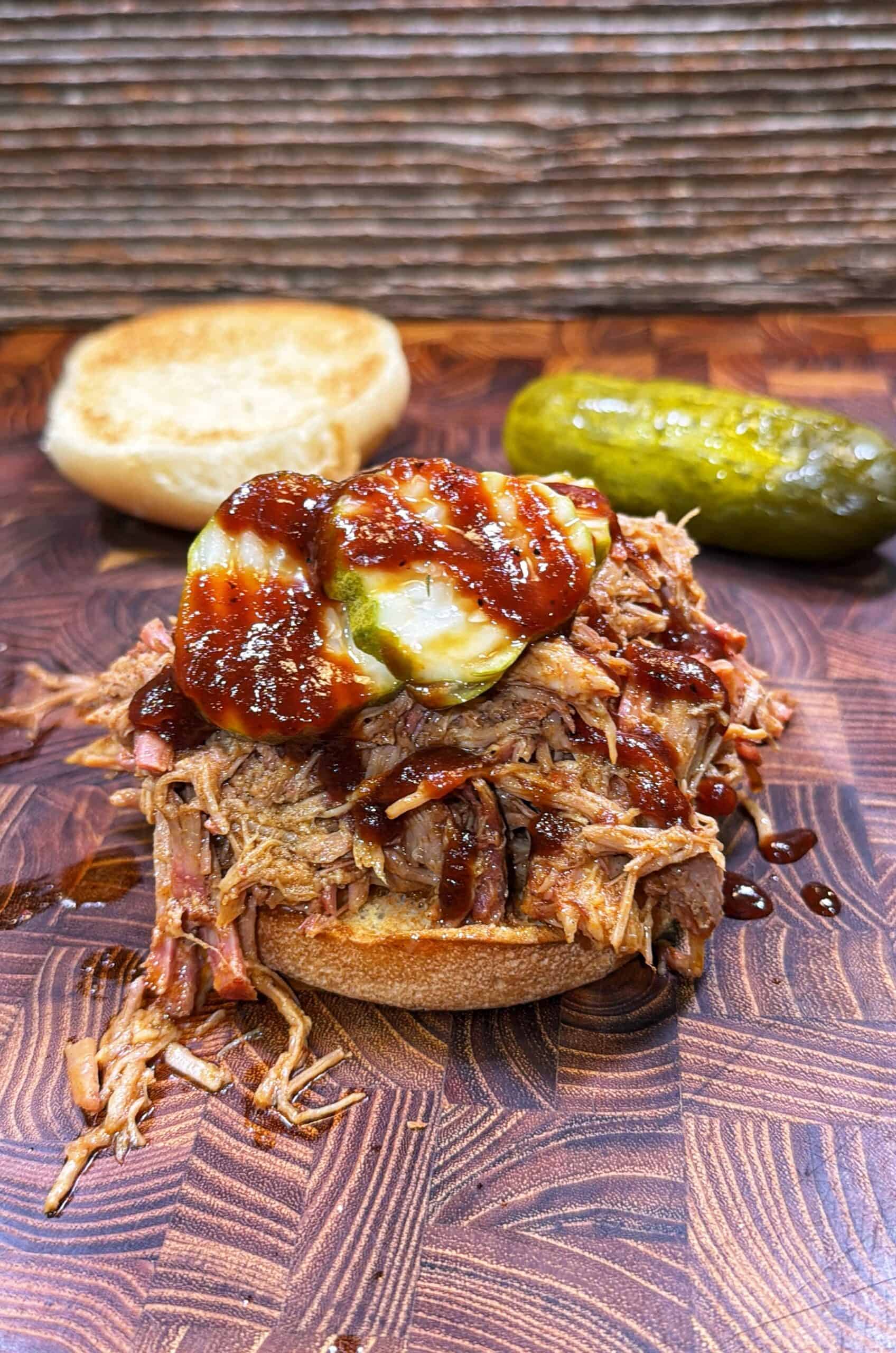 Pulled pork sandwich with barbecue sauce and pickles on a wooden surface, with the top bun off to the side and a whole pickle in the background.