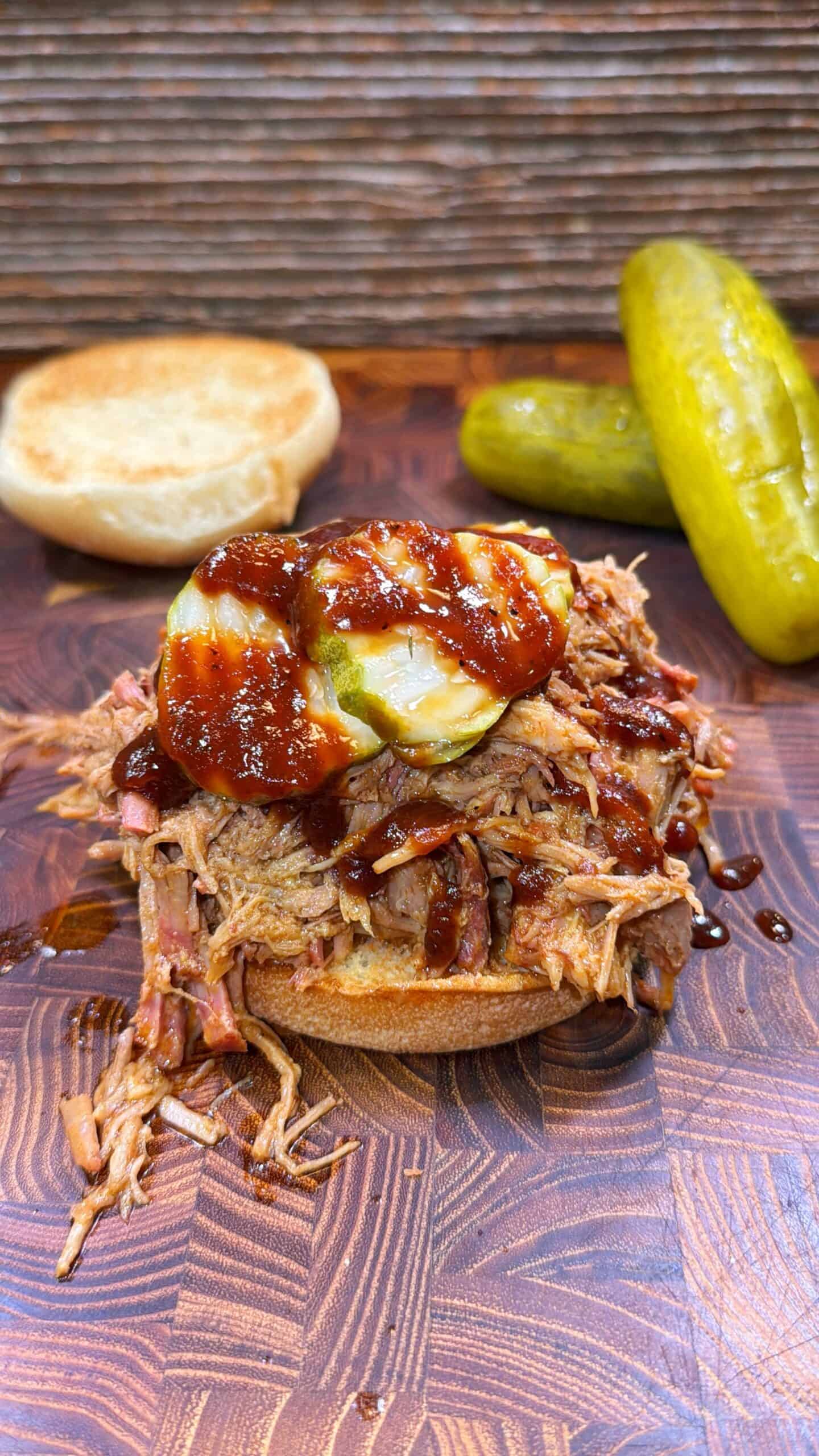 Pulled pork sandwich topped with pickles and barbecue sauce on a wooden board, with a split bun and two whole pickles in the background.