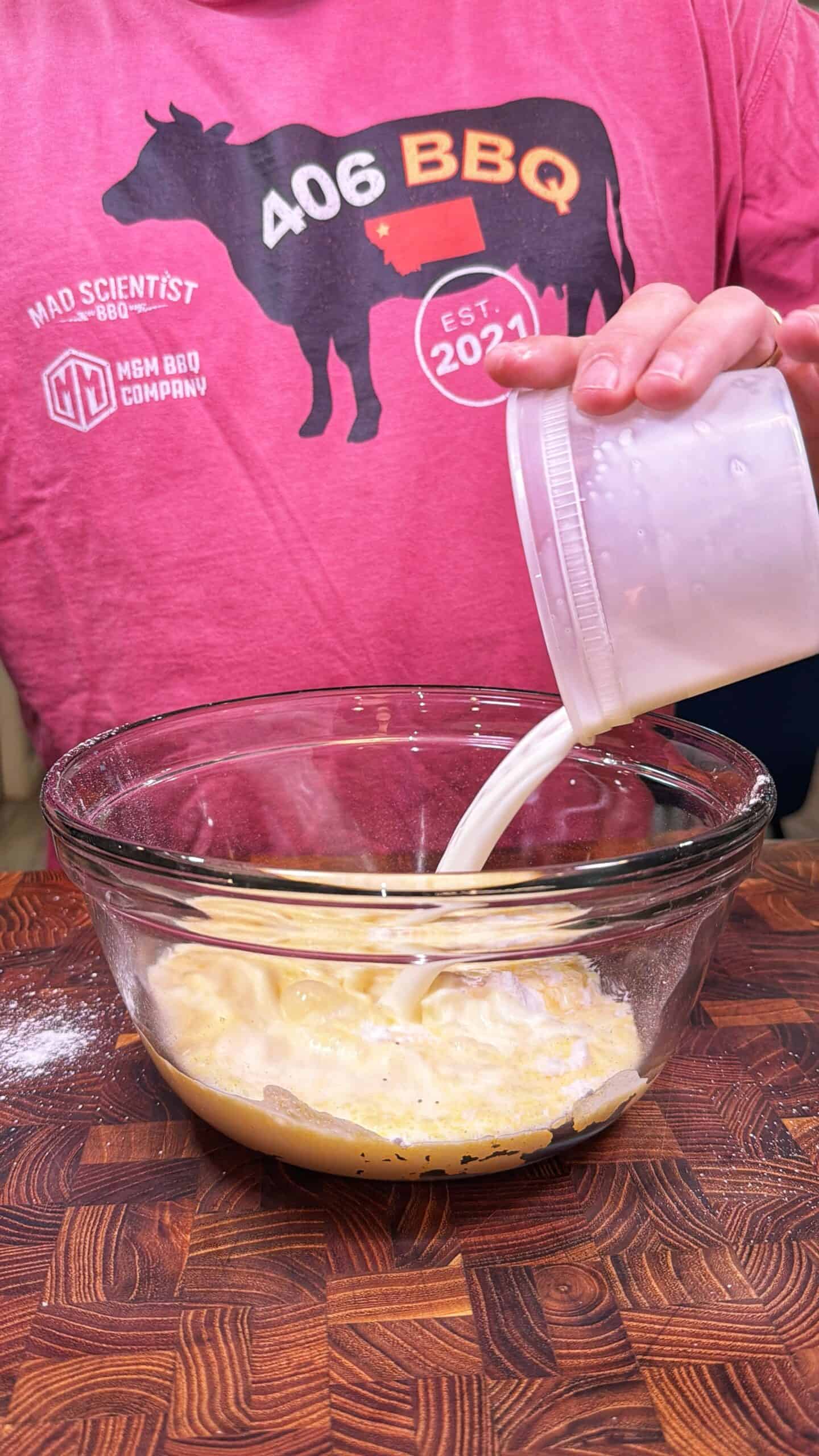 A person in a pink “406 BBQ” shirt pours milk from a plastic container into a glass bowl with a beaten mixture on a wooden surface.