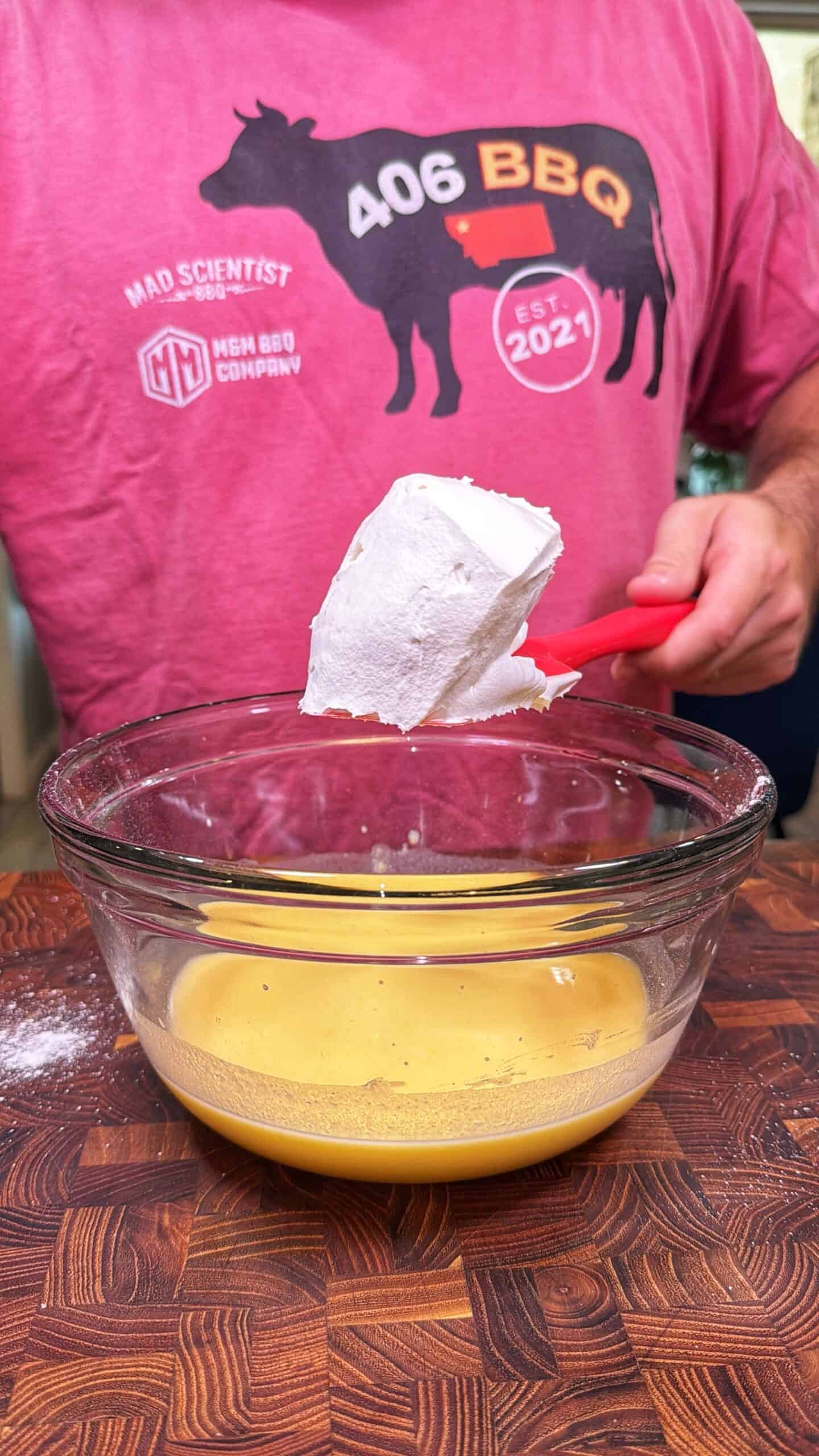 A person in a pink BBQ shirt adds a scoop of white flour to a glass bowl with a yellow batter, on a wooden surface.