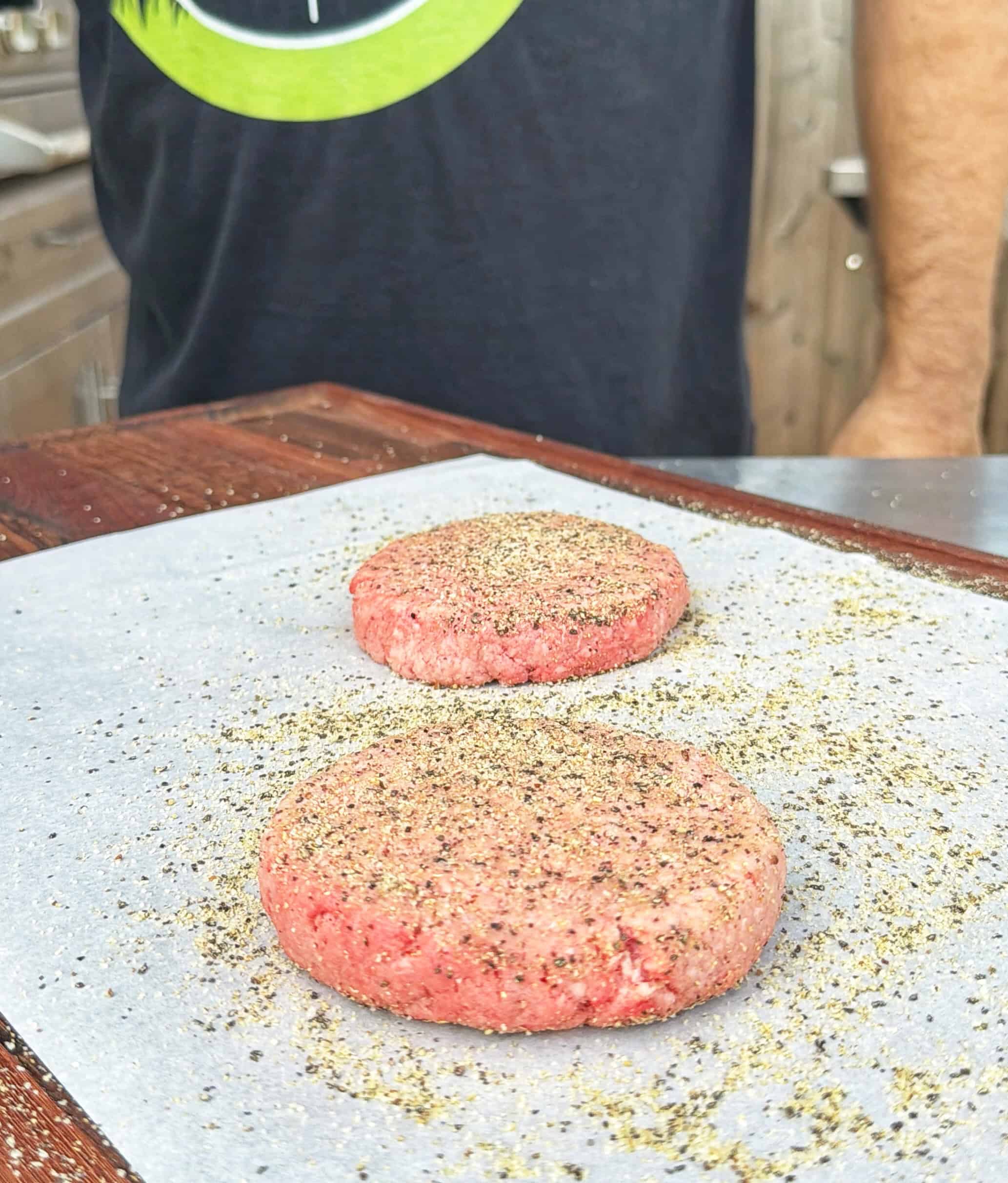 Two raw hamburger patties, seasoned with black pepper, sit on parchment paper atop a wooden surface. A person wearing a dark t-shirt stands behind the patties.