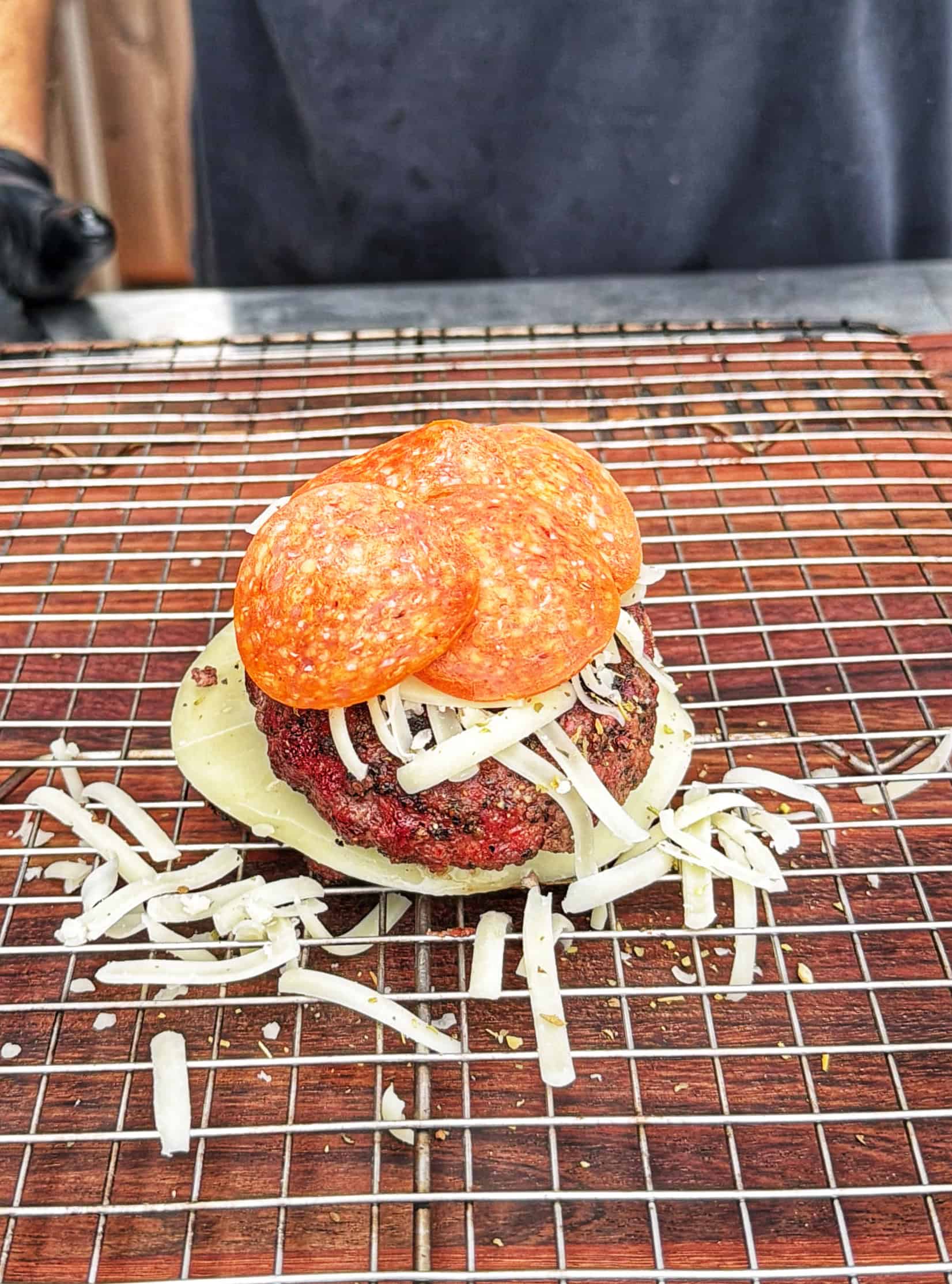 A burger patty topped with shredded cheese, a slice of cheese, and three slices of pepperoni, sits on a wire rack over a wooden surface. A person in dark clothing is visible in the background.