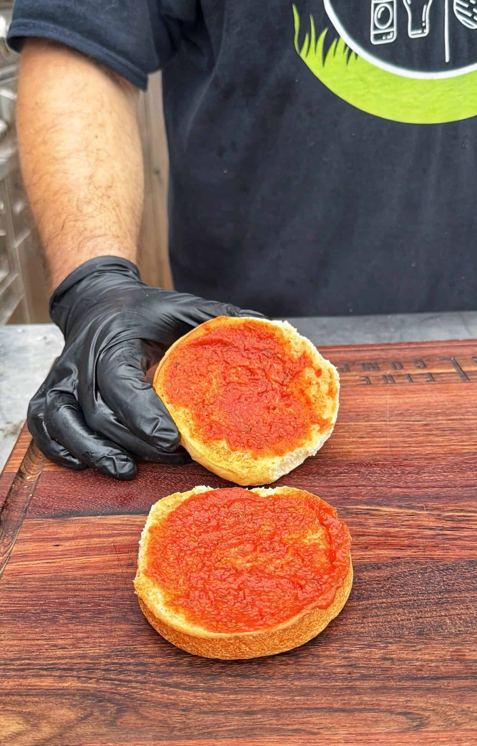 A person wearing a black glove holds a sandwich bun, both halves spread with tomato sauce, on a wooden <a href=