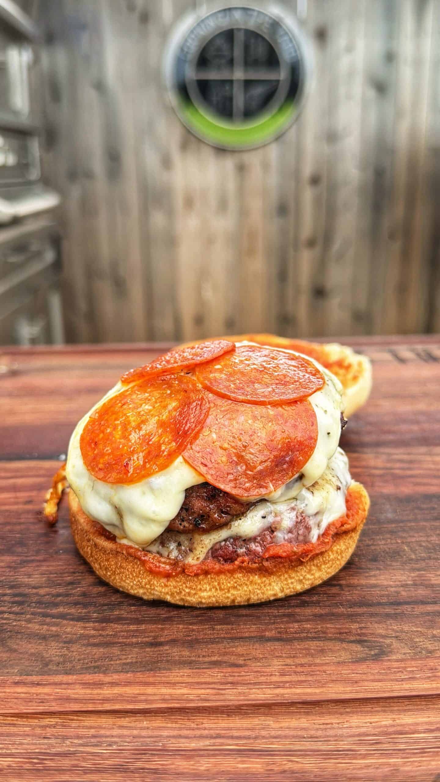 An open hamburger bun topped with melted cheese, pepperoni slices, and a beef patty sits on a wooden surface, with a blurred wooden fence and a round window in the background.