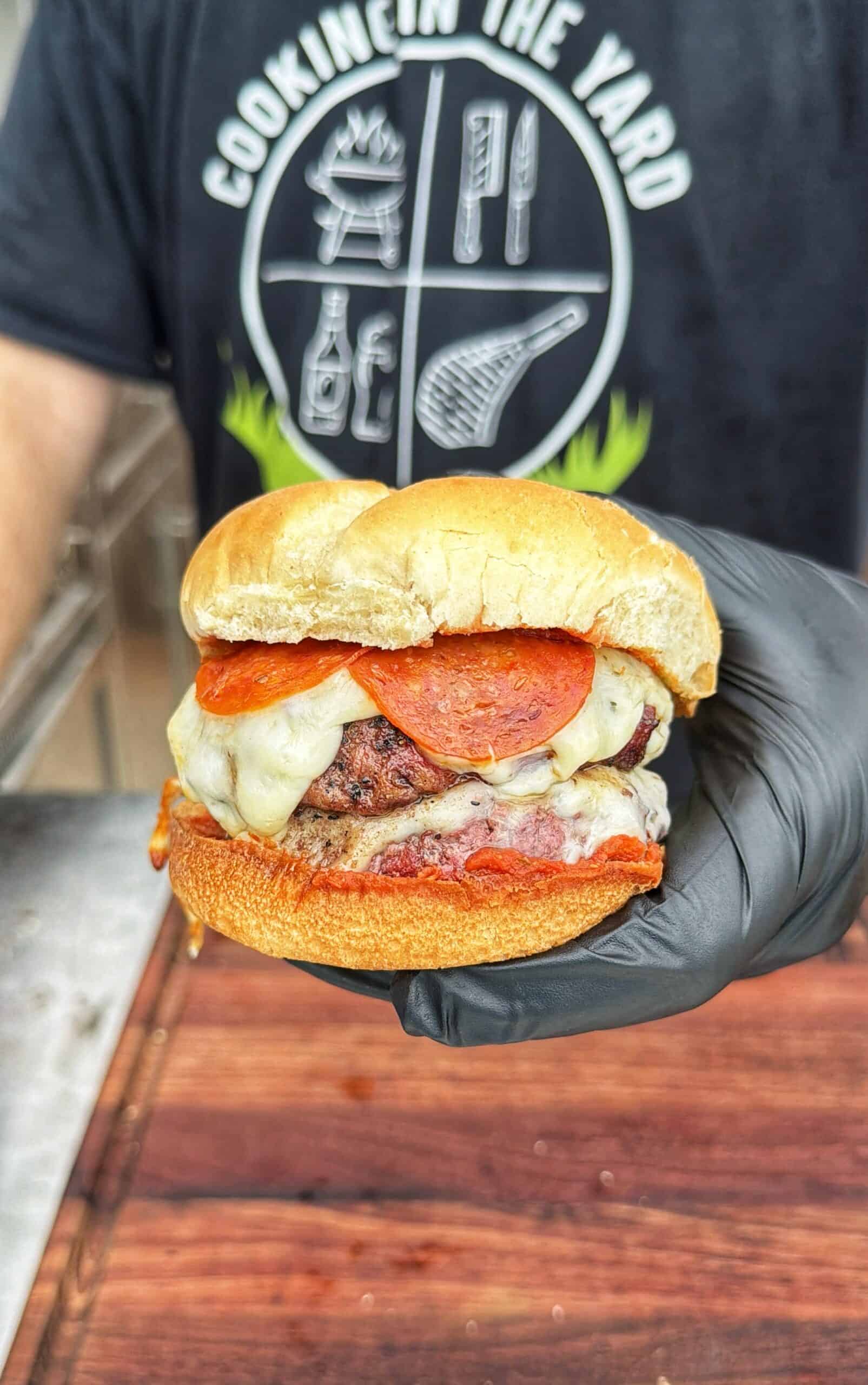 A gloved hand holds a cheeseburger topped with pepperoni and melted cheese. The person wears a Cookin in the Yard shirt with cooking utensils graphic in the background.