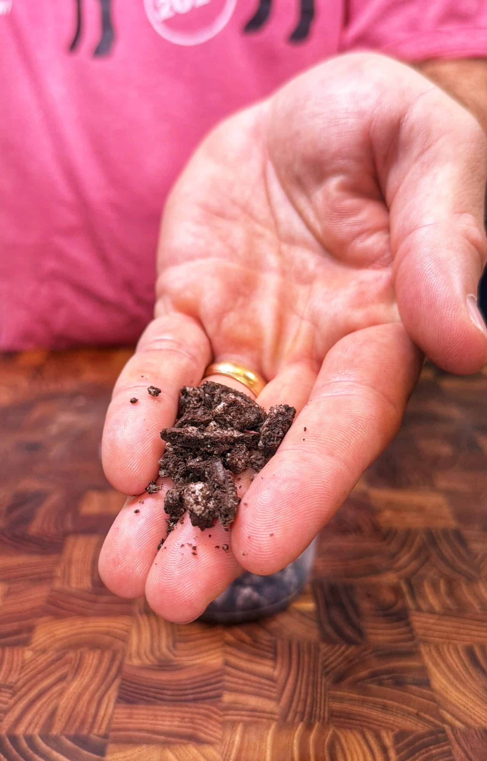 A hand holds crumbled chocolate cookie pieces over a wooden surface. The person is wearing a pink shirt and a gold ring.