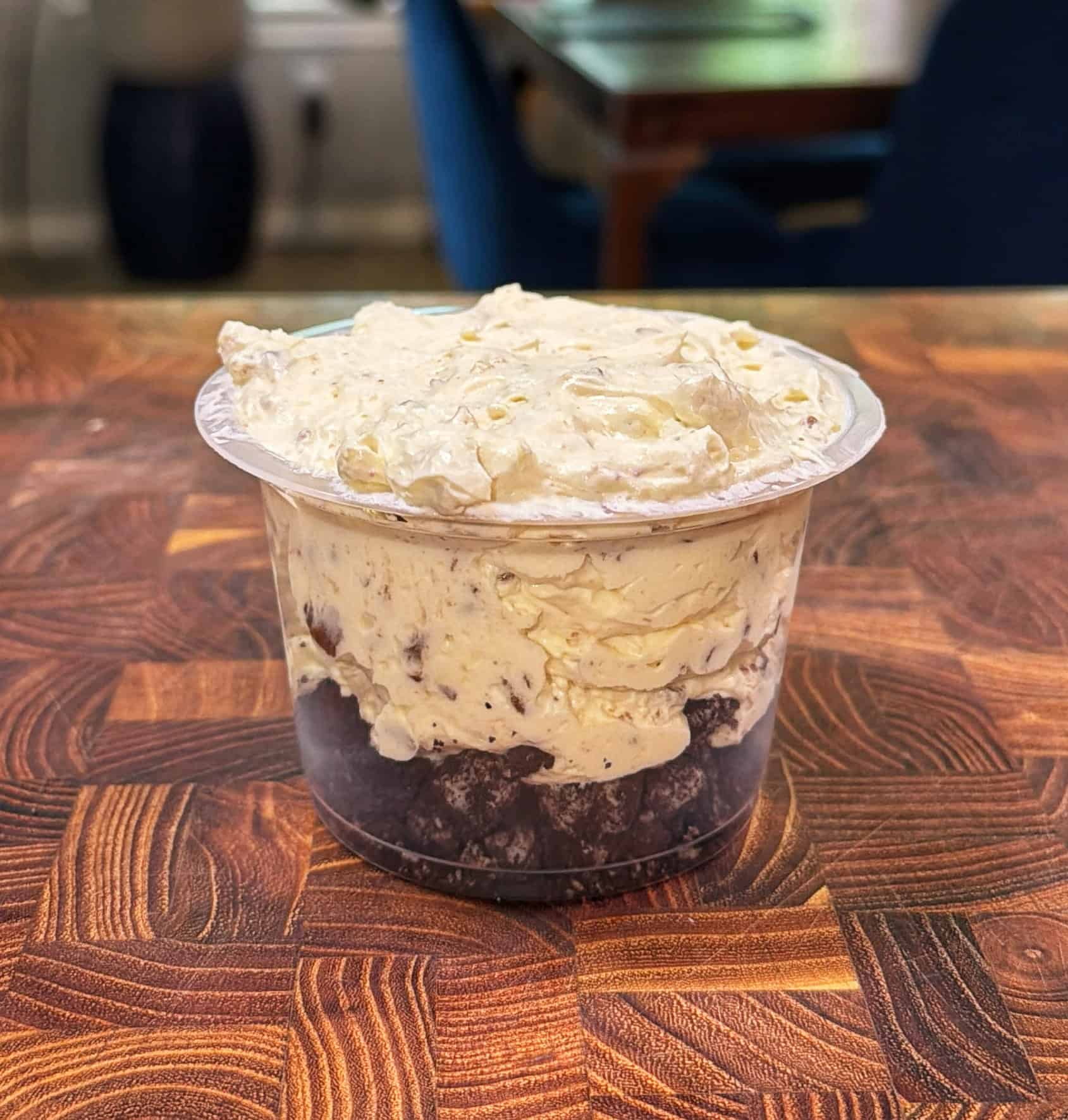 A clear plastic cup filled with crumbled chocolate brownie pieces at the bottom and topped with a thick layer of creamy, whipped dessert, sitting on a wooden patterned surface.