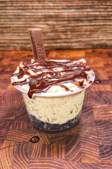 A dessert cup with chocolate drizzle, creamy filling, a chocolate base, and a Kit Kat piece on top, placed on a wooden surface.