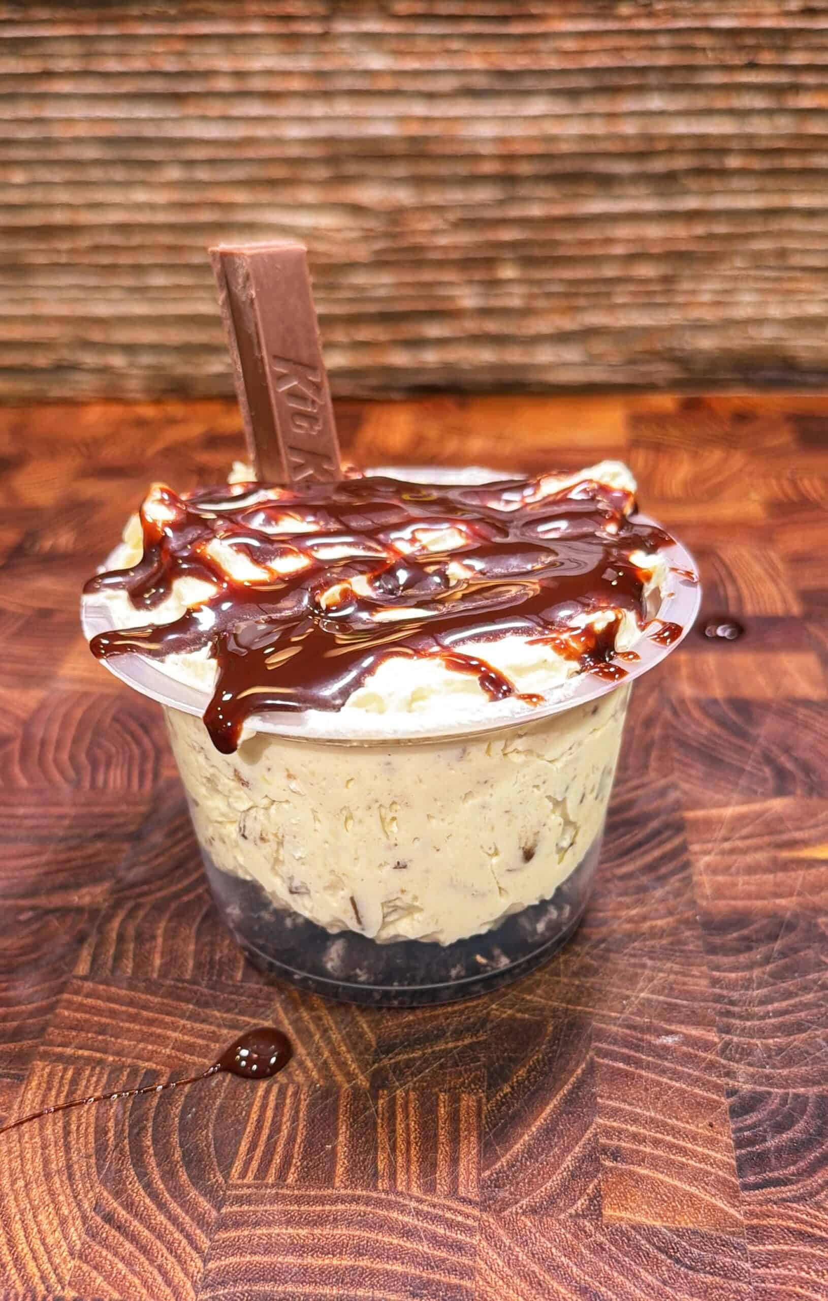 A dessert cup with chocolate drizzle, creamy filling, a chocolate base, and a Kit Kat piece on top, placed on a wooden surface.