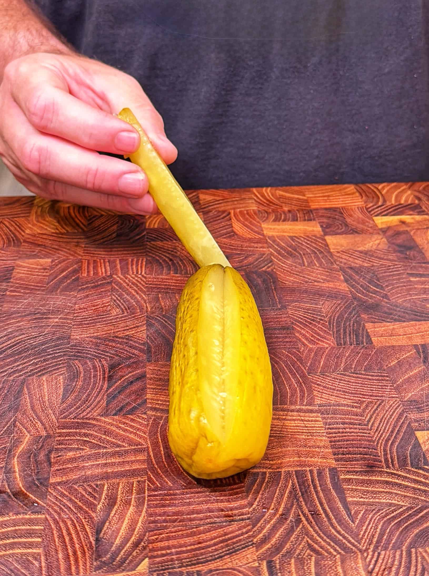 A hand inserts a pickle spear into a slit made in a whole pickle on a wooden <a href=