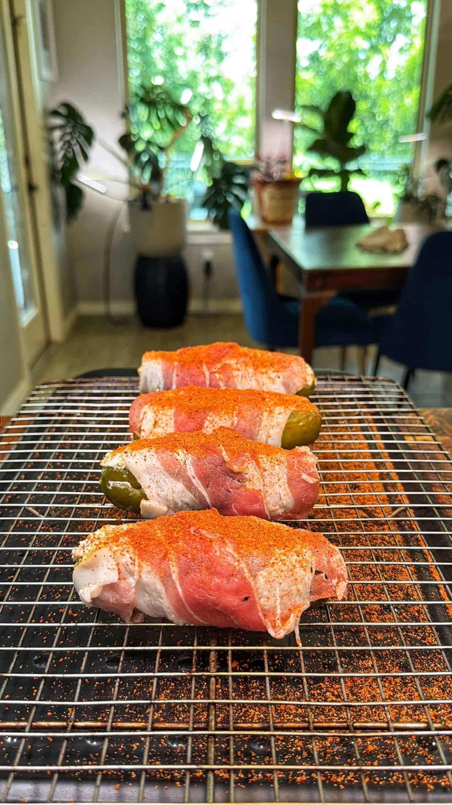 Four bacon-wrapped jalapeños sprinkled with seasoning sit on a wire rack, with a bright, plant-filled dining room in the background.